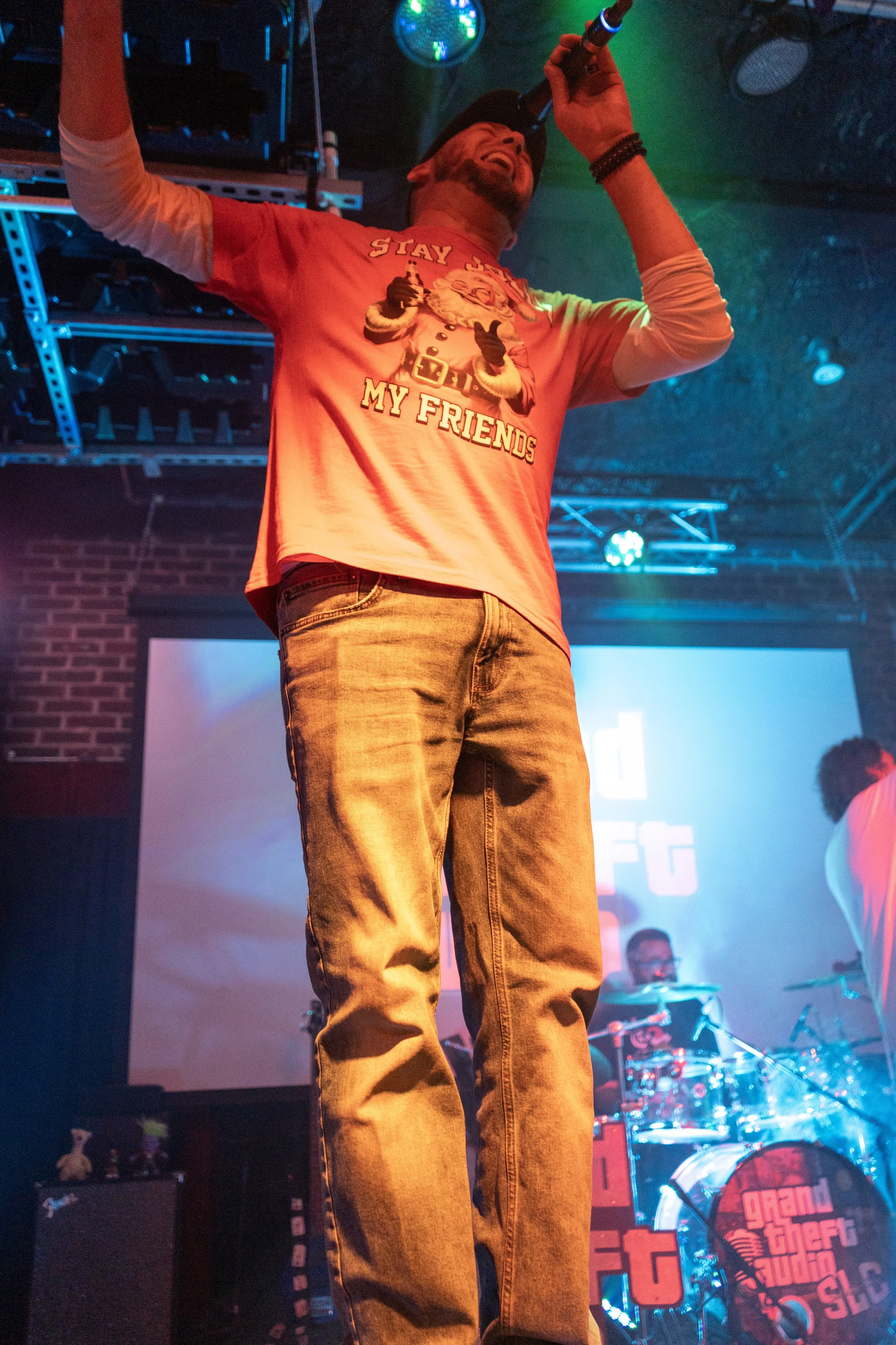 A man singing into a microphone on stage at a concert or club. He's wearing a red T-shirt with a graphic and text, jeans, and a black wristband. The background shows stage equipment and colorful lighting.