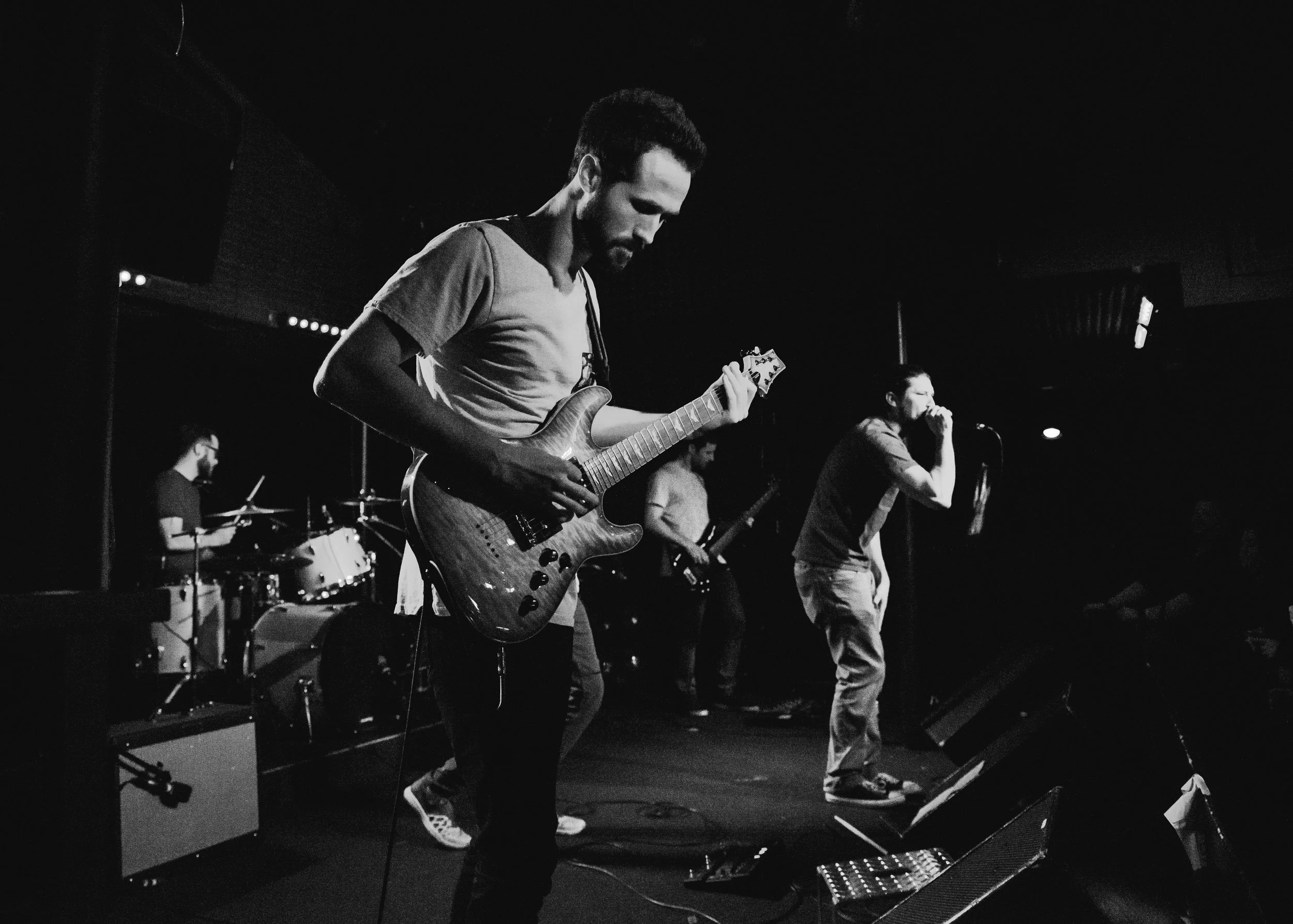 Black and white photo of a band performing on stage, with a guitarist in the foreground and a singer holding a microphone in the background.
