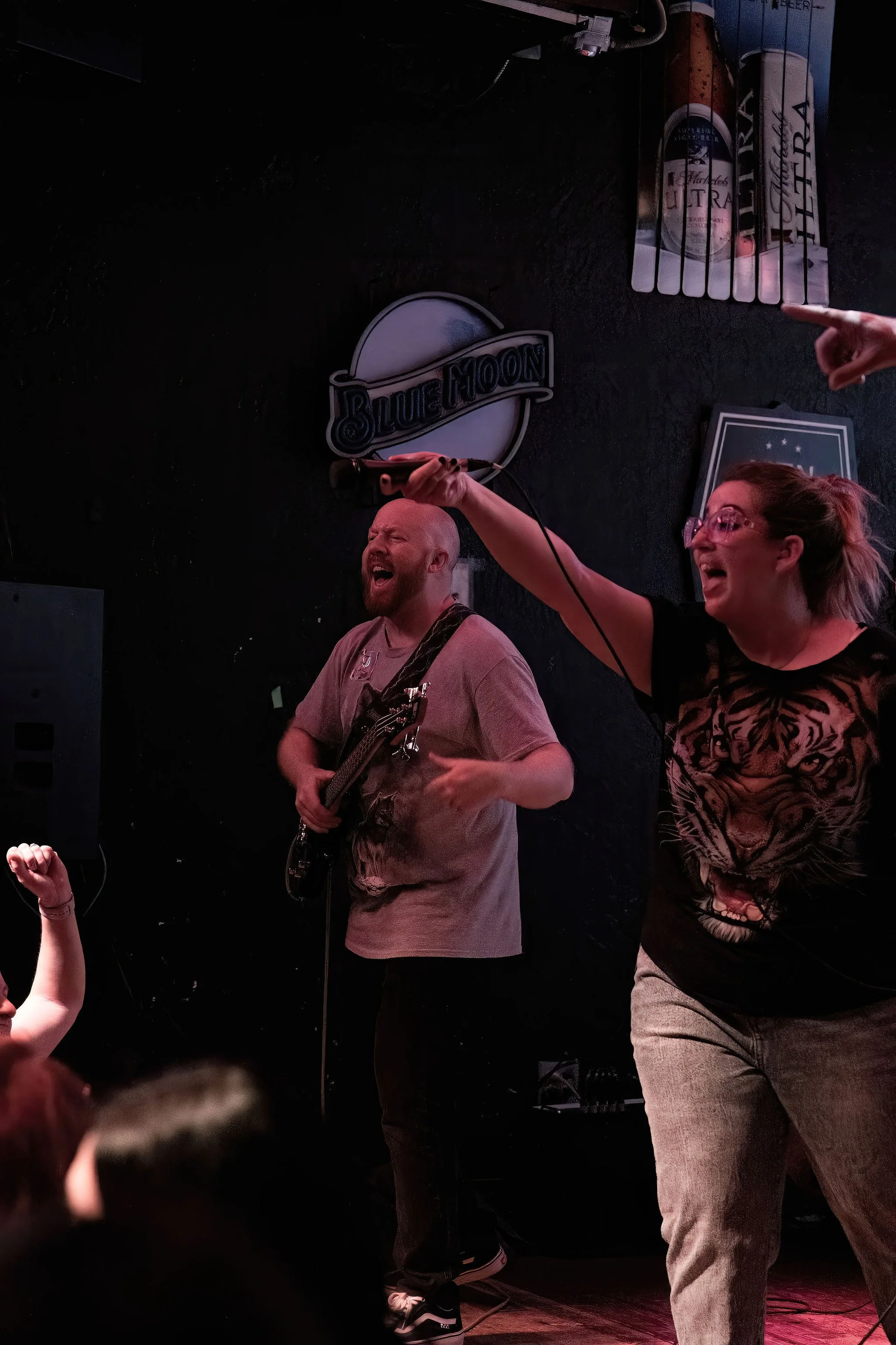 A woman with glasses and a shirt with a tiger print singing into a microphone, accompanied by a man with a guitar, on a stage with a Blue Moon sign and beer advertisement posters in the background. Audience members are visible in the foreground.