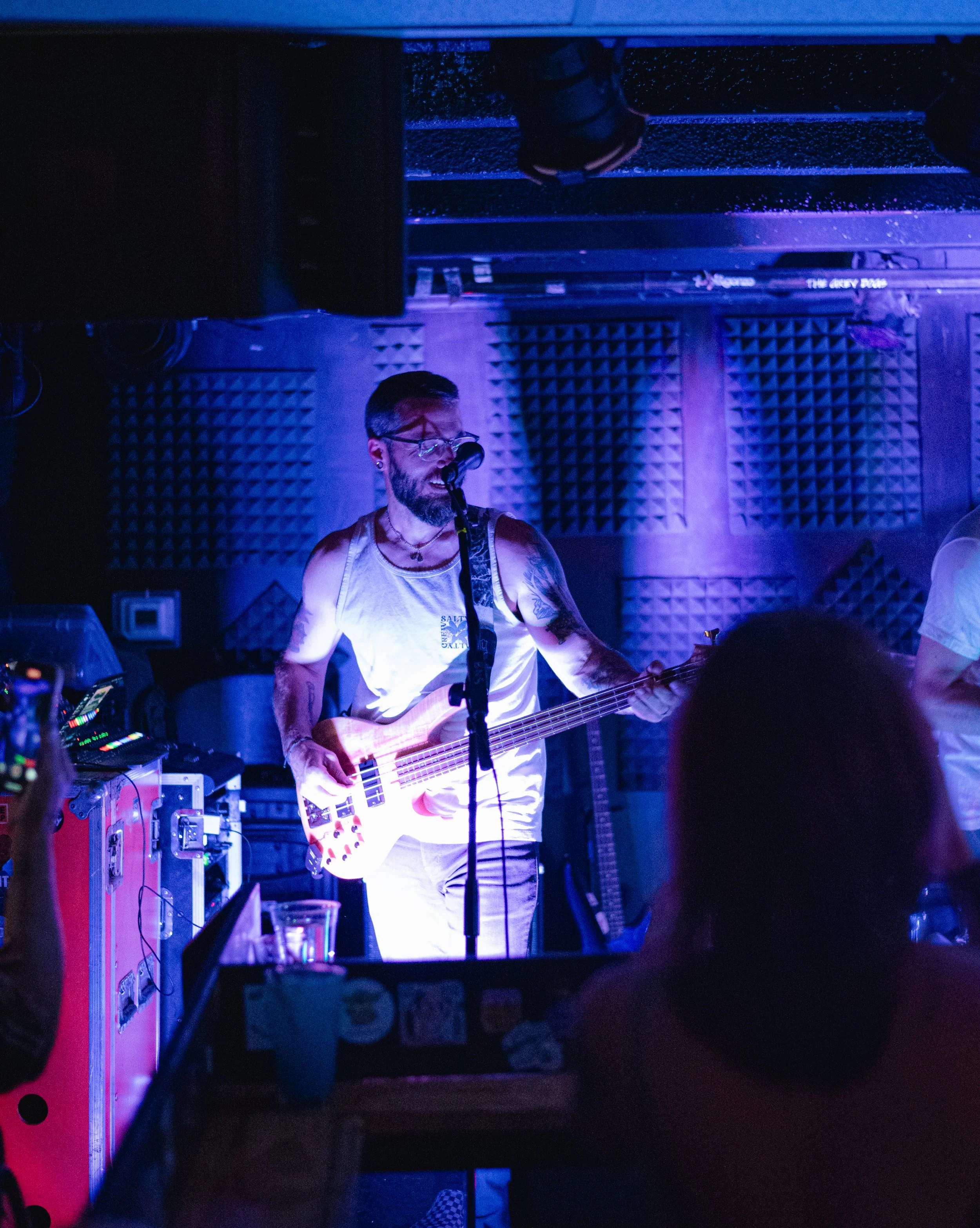 A man with glasses and a beard playing a bass guitar on stage with purple lighting, surrounded by sound equipment and foam acoustic panels.