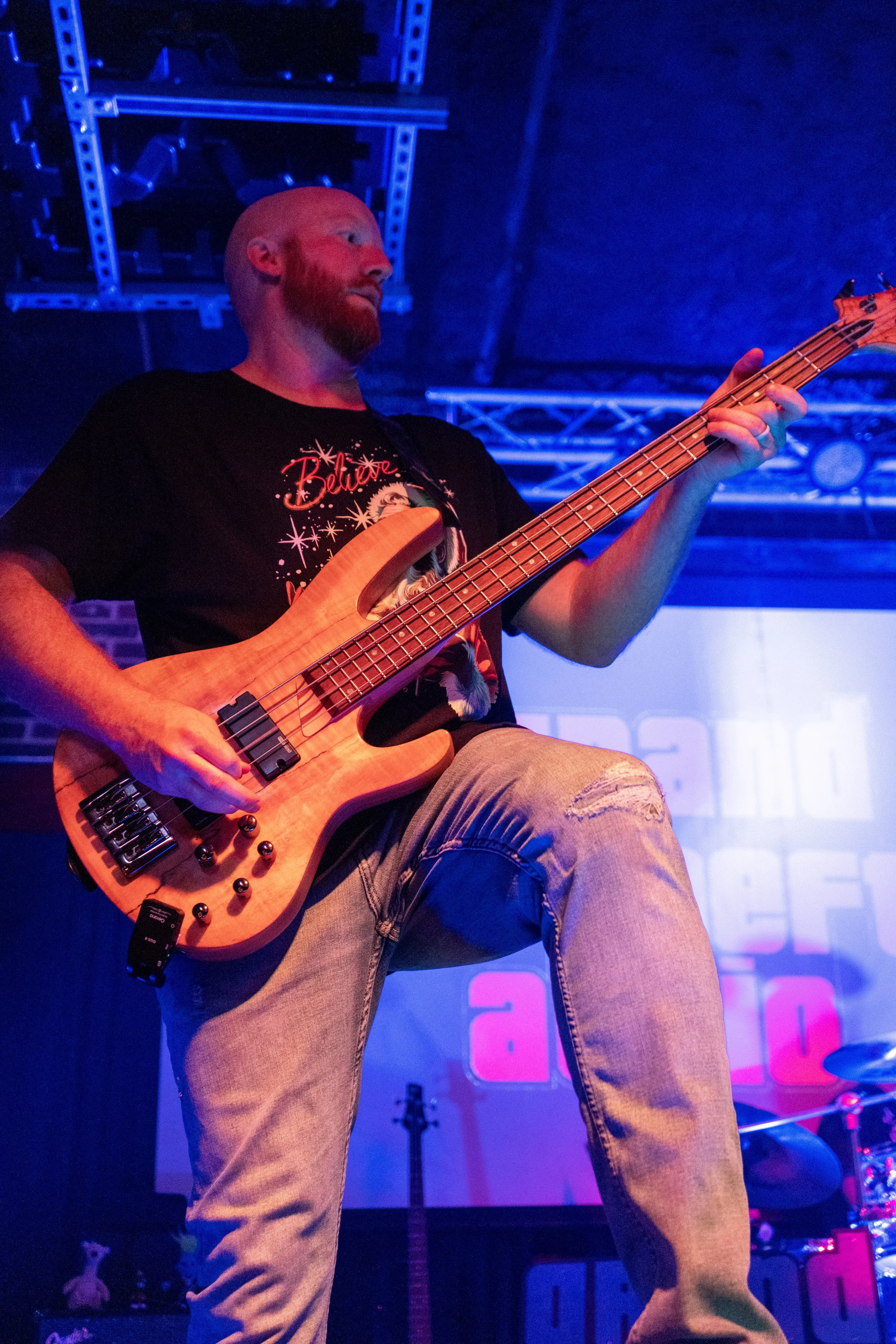 A man playing an electric bass guitar on stage with blue and purple lighting, wearing a black t-shirt and ripped jeans.