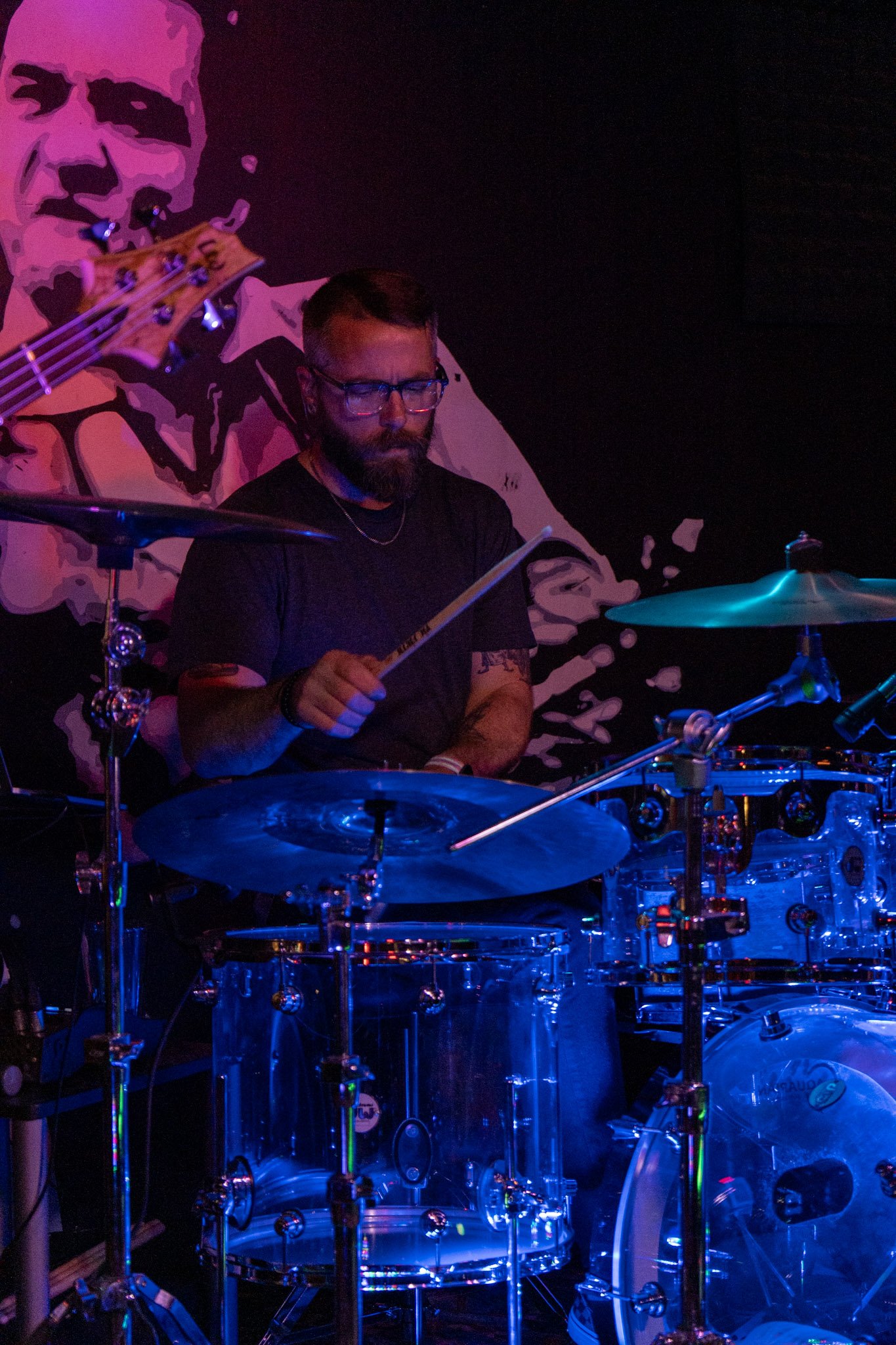Nightclub scene with a man playing drums and a mural of a man holding a guitar in the background, illuminated with blue and purple lighting.