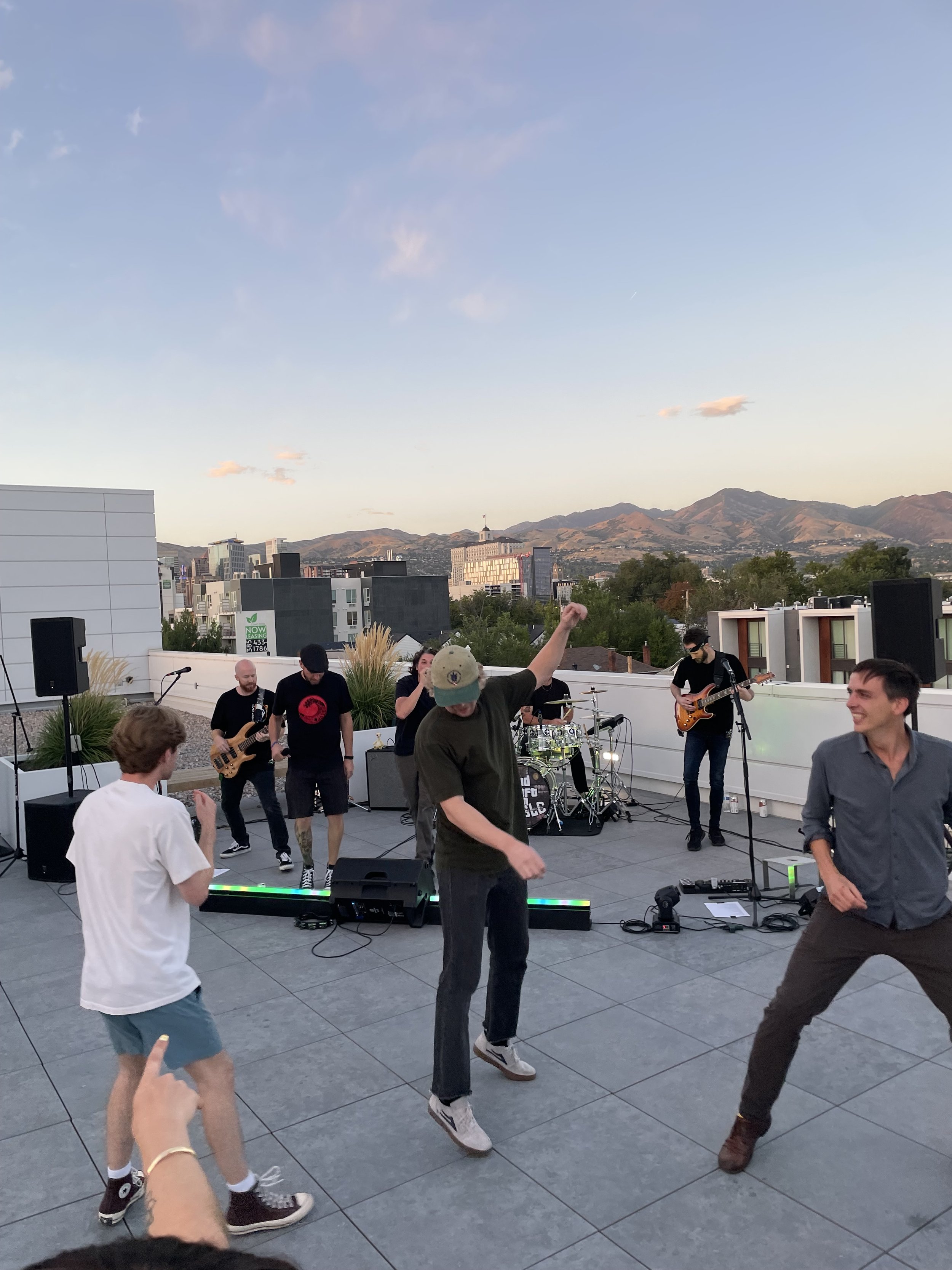 A group of people dancing and enjoying music at an outdoor rooftop concert during sunset, with city buildings and mountains in the background.