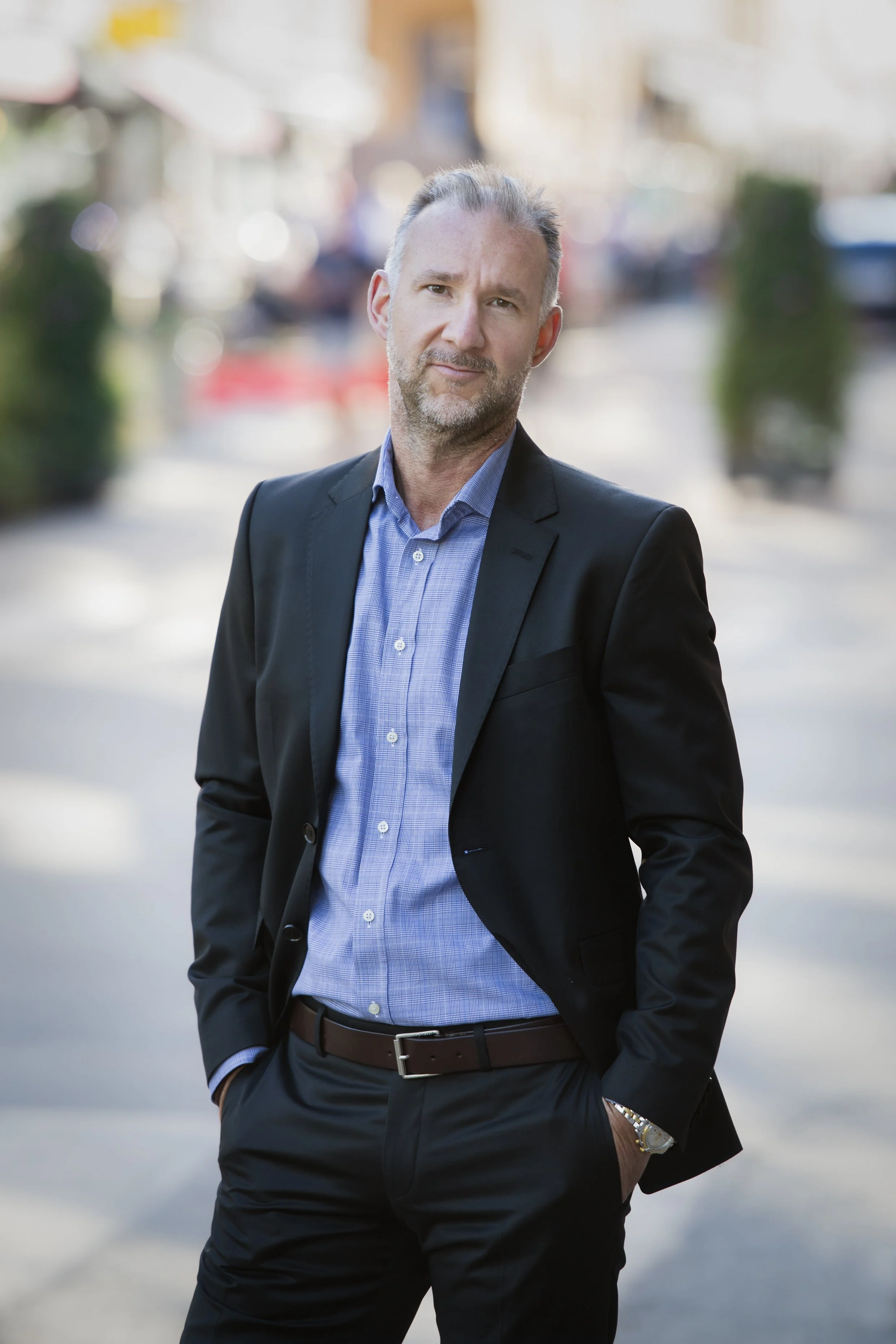 A middle-aged man in a business suit with a blue shirt, standing outdoors in a city street with blurred buildings and vehicles in the background.