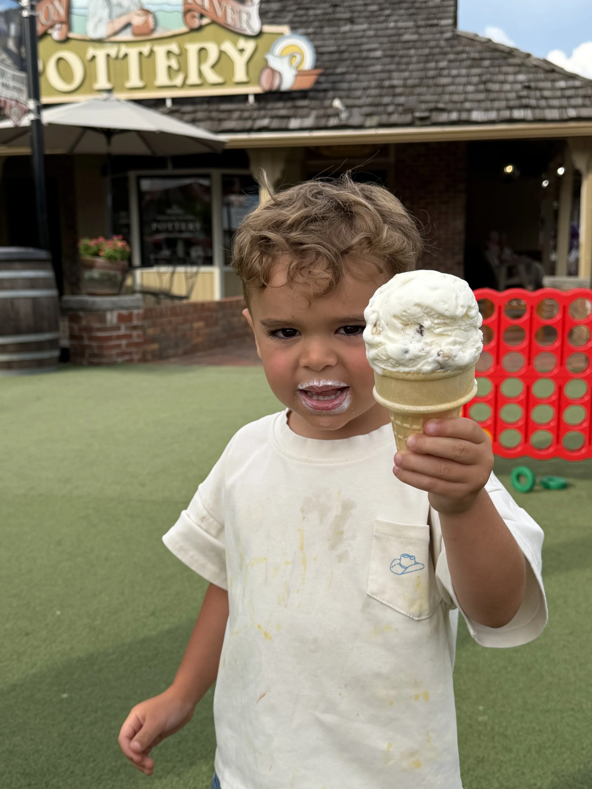 A young boy is holding an ice cream cone, standing outdoors near a building with a sign that reads 'Lemon Lottery'. The boy is wearing a light-colored t-shirt with some stains, and has curly light brown hair. The background shows green artificial gra