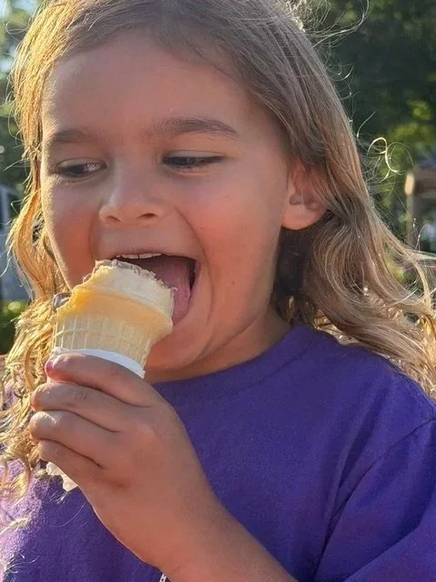 A young girl with light brown hair wearing a purple shirt is licking vanilla ice cream from a cone outdoors during daylight.