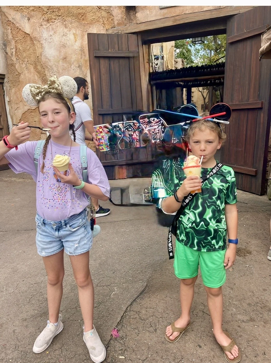 Two children, a girl and a boy, are enjoying ice cream cones outside near a rustic wooden and stone building. The girl has braided hair, a purple T-shirt with colorful designs, denim shorts, and Mickey Mouse ears. The boy has curly hair, a green camo