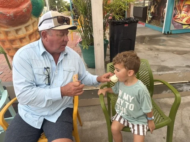 An older man and a young boy sharing a cone of ice cream at an outdoor seating area. The man is sitting on a yellow chair, wearing a light blue shirt, a white cap, and sunglasses on his head. The boy is sitting on a green chair, wearing a gray T-shirt and striped shorts, licking the ice cream.