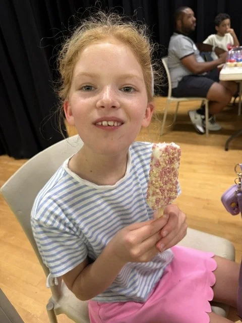 A young girl with red hair and freckles sitting on a chair, holding a pink and white ice cream bar covered in sprinkles. She is smiling slightly and wearing a blue and white striped shirt and pink shorts. In the background, two boys are sitting at a 
