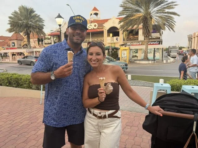 A man and woman standing outdoors in a shopping or dining area with palm trees, holding ice cream cones and smiling at the camera. The man is wearing a blue patterned shirt, black shorts, and a baseball cap that says 'Cowboy Golf'. The woman is weari