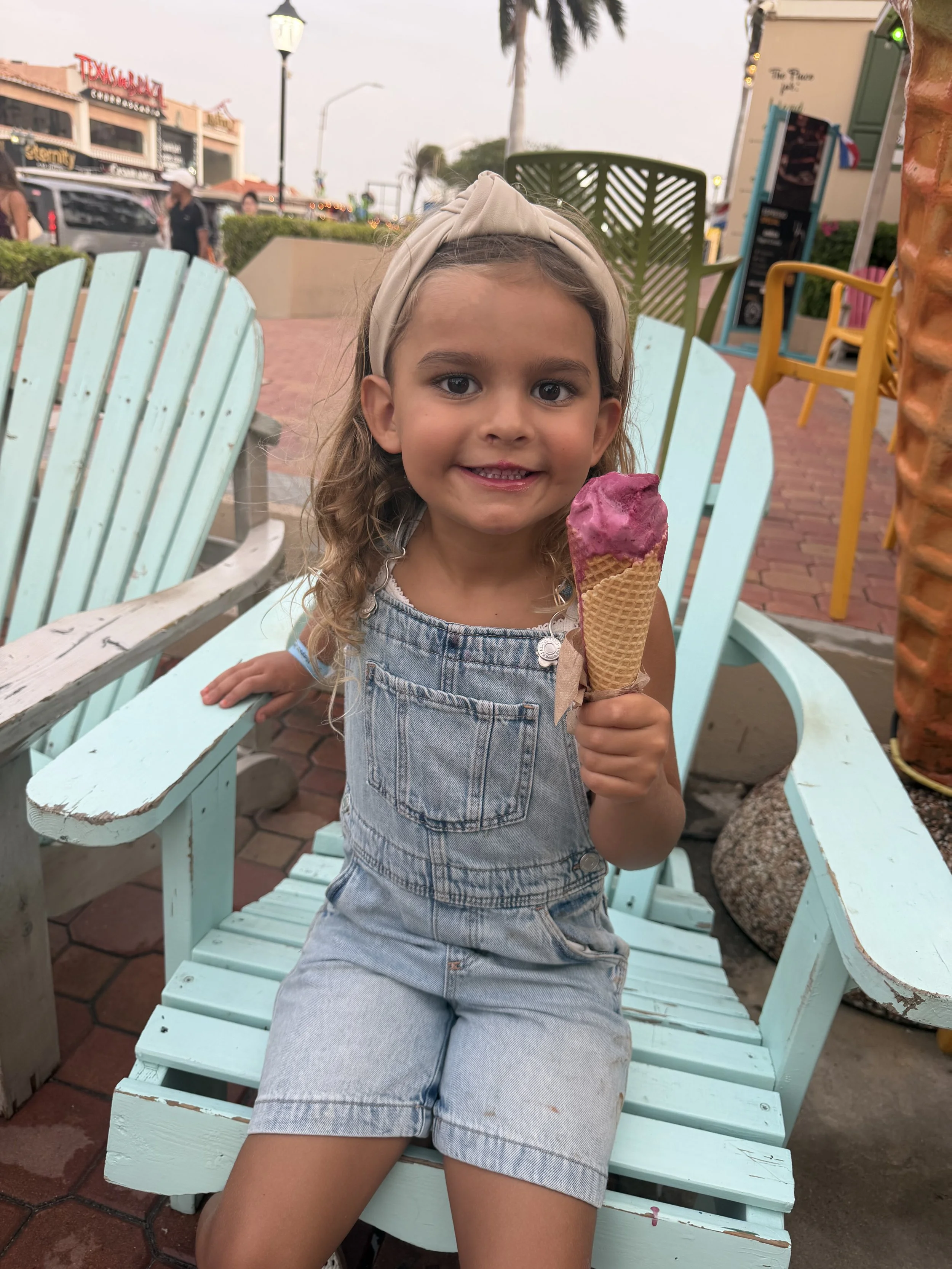A young girl with curly hair wearing a beige headband and denim overalls, sitting on a light blue bench outdoors, holding a pink ice cream cone, with palm trees, buildings, and a cloudy sky in the background.