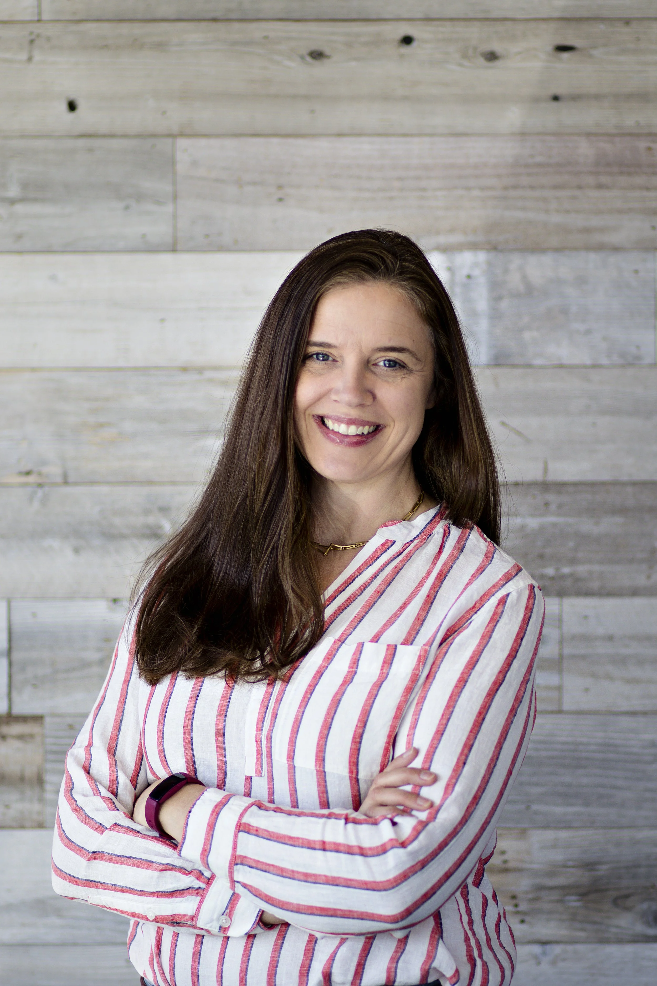 A woman with long brown hair smiling, wearing a striped shirt, standing against a wooden wall background.