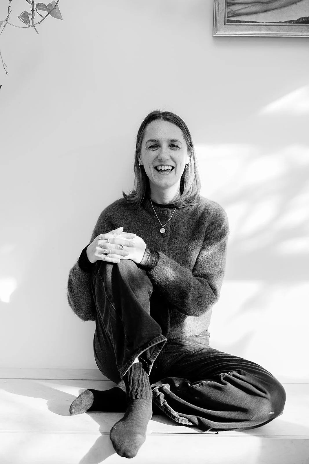 Amy Melson Somatic Coach in London sitting cross-legged indoors in the shadows of leaves from outdoors.