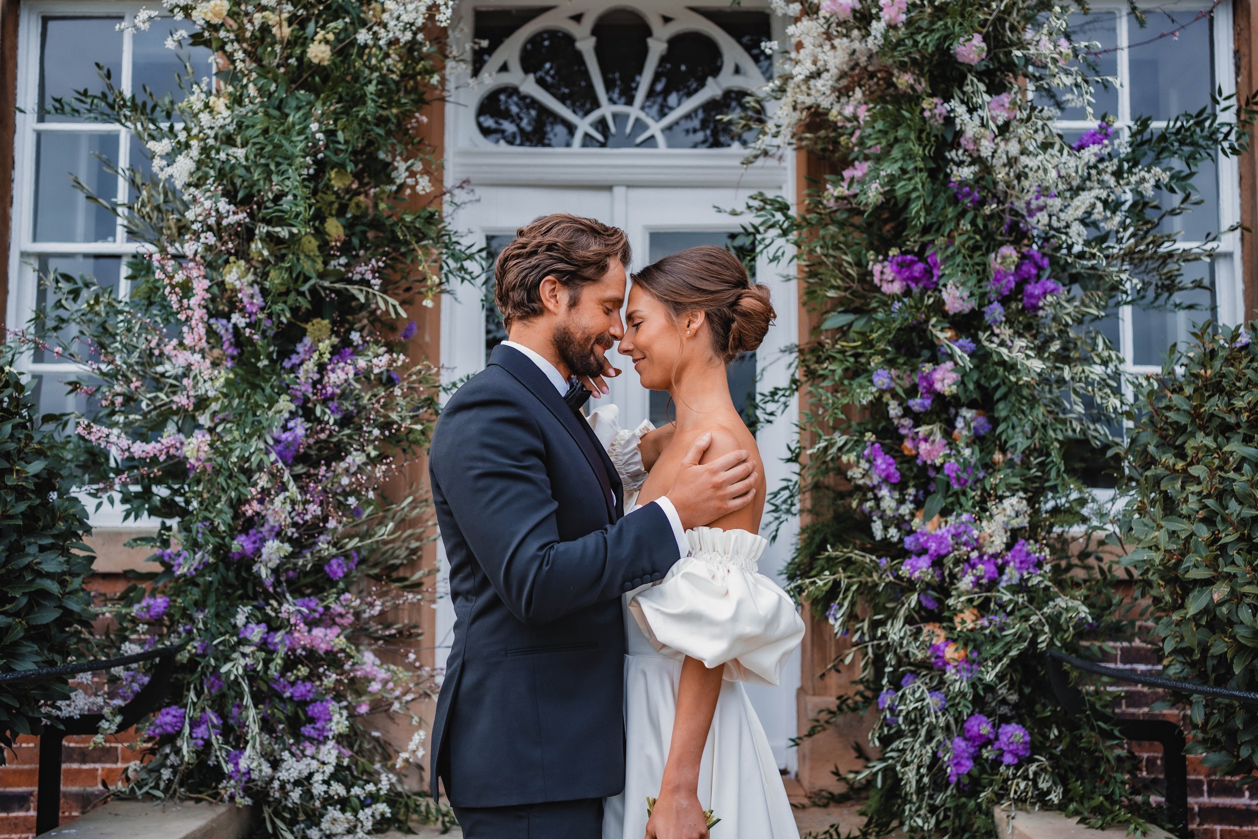 A couple dressed in wedding attire sharing a moment in front of a floral arrangement and a white door with decorative glass.
