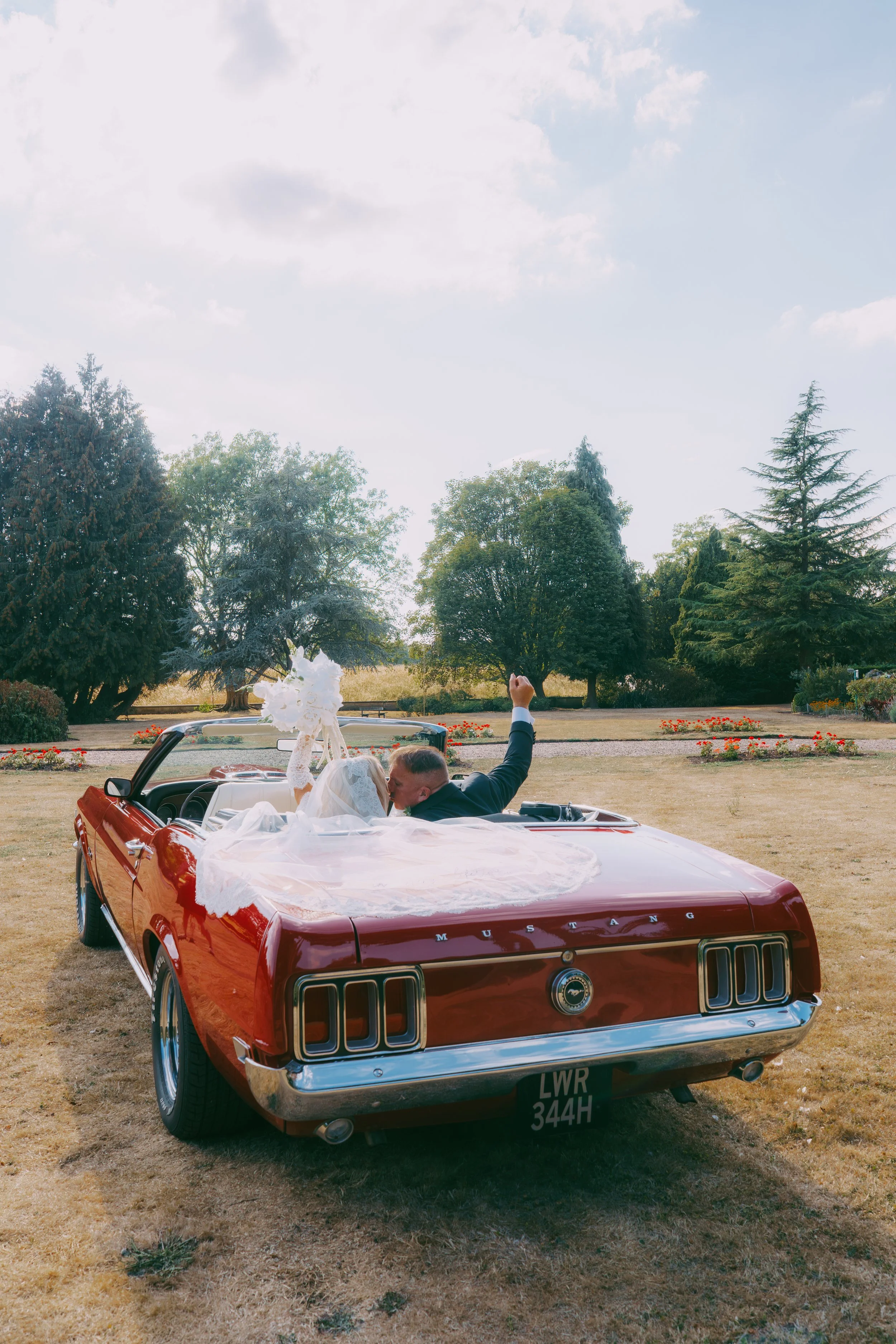 A couple sitting in the back of a red vintage Ford Mustang convertible, which is decorated for a wedding, with a scenic park in the background and trees lining the horizon.