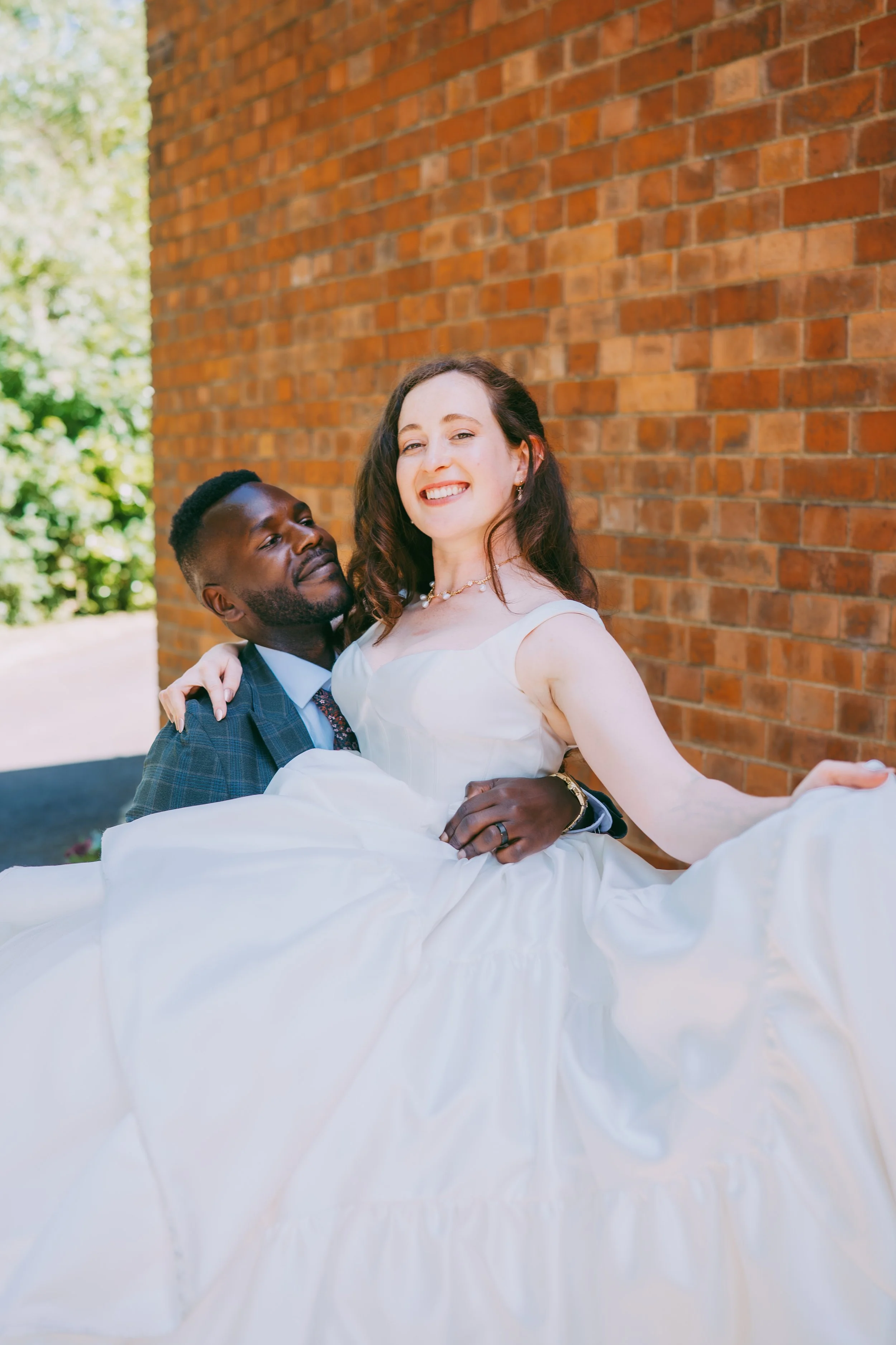 A bride and groom smiling and posing outdoors against a brick wall, with greenery in the background. The bride is wearing a white wedding dress and the groom is in a dark suit.