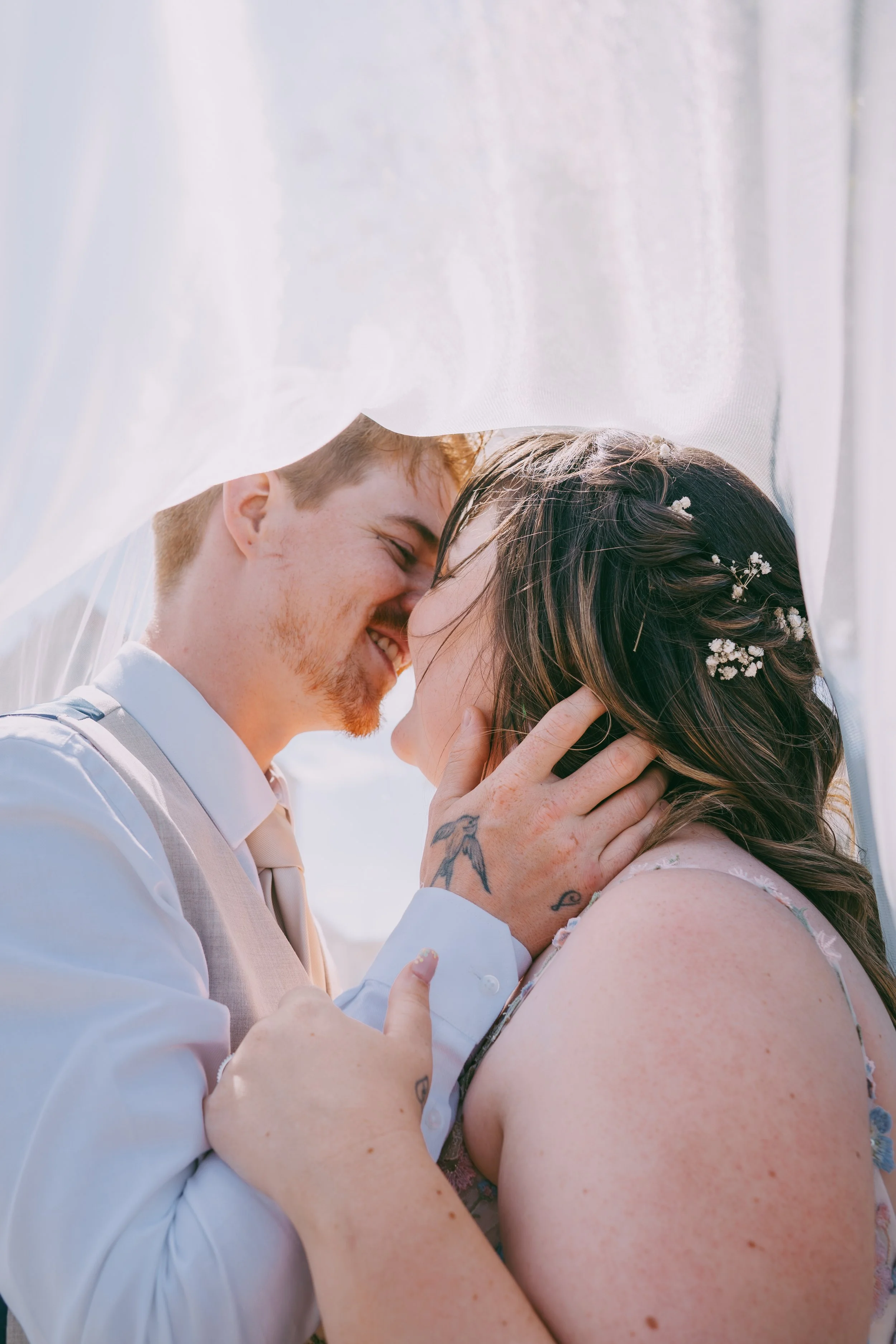 A couple on their wedding day, smiling and touching foreheads, with the bride's face partly obscured by a flowing white fabric. The groom has reddish hair and a beard, and the bride has brown hair with small white flowers tucked in. They appear very 