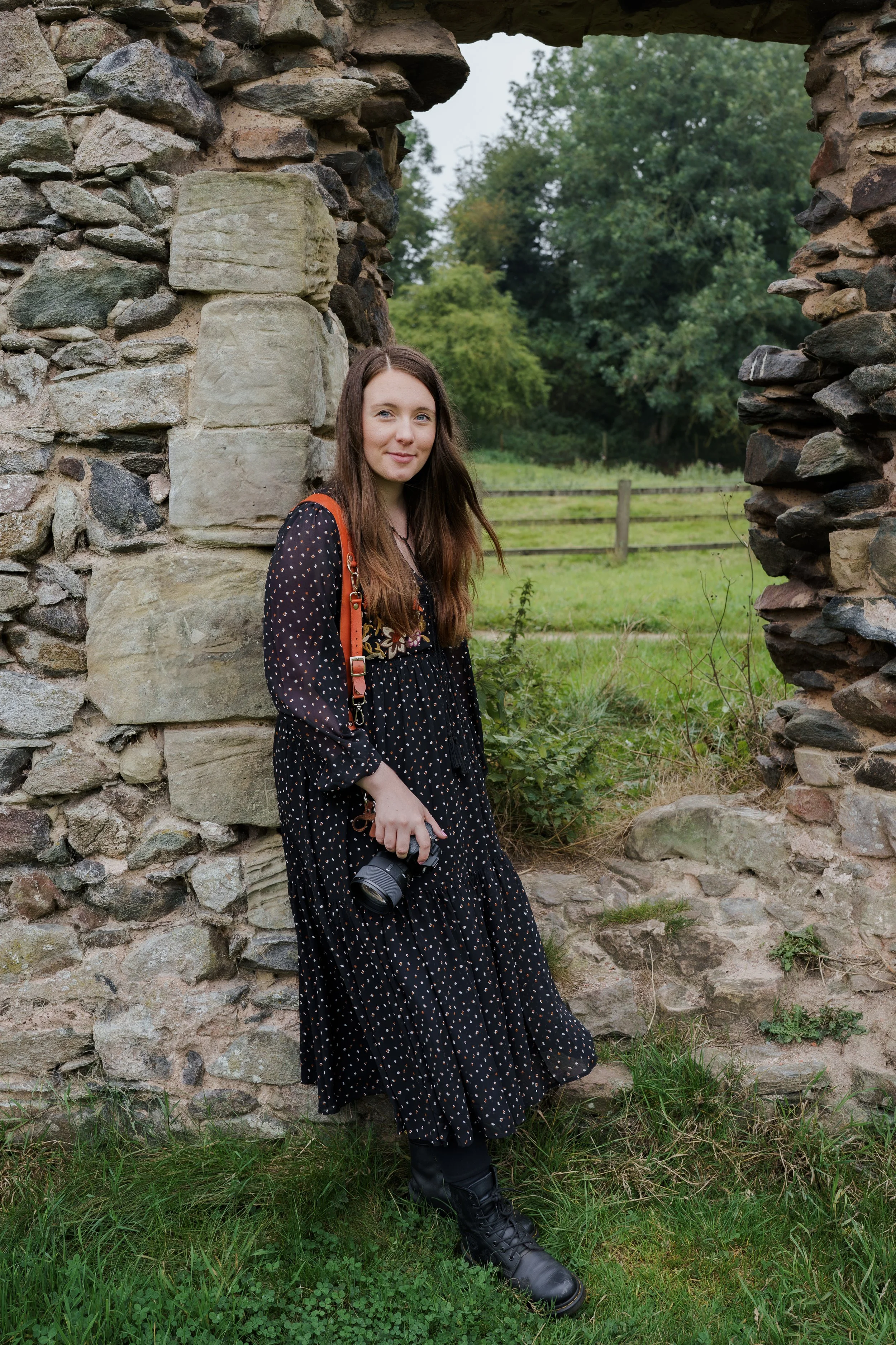 A young woman with long brown hair wearing a black dress with white polka dots, black boots, and carrying a camera, standing beside a stone wall with a grassy field and trees in the background.