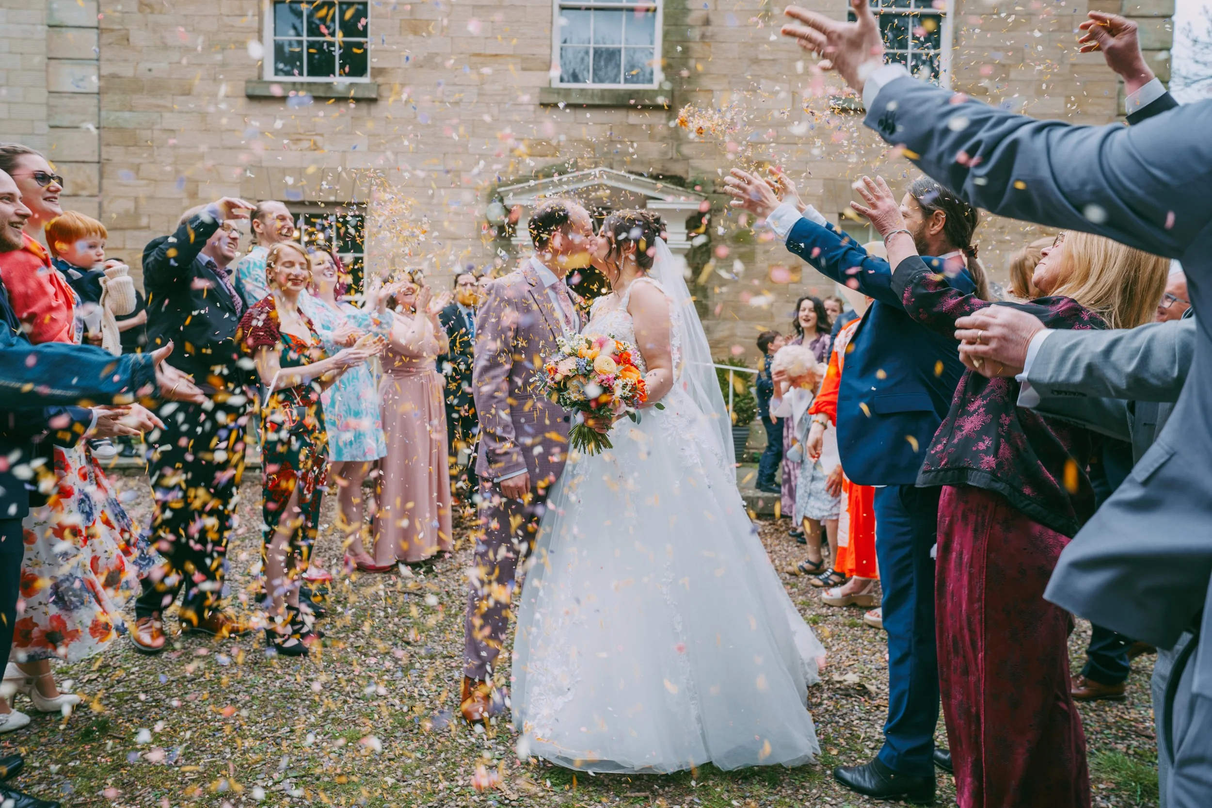 A wedding celebration with a bride and groom kissing, surrounded by happy guests throwing colorful confetti outside a stone building.