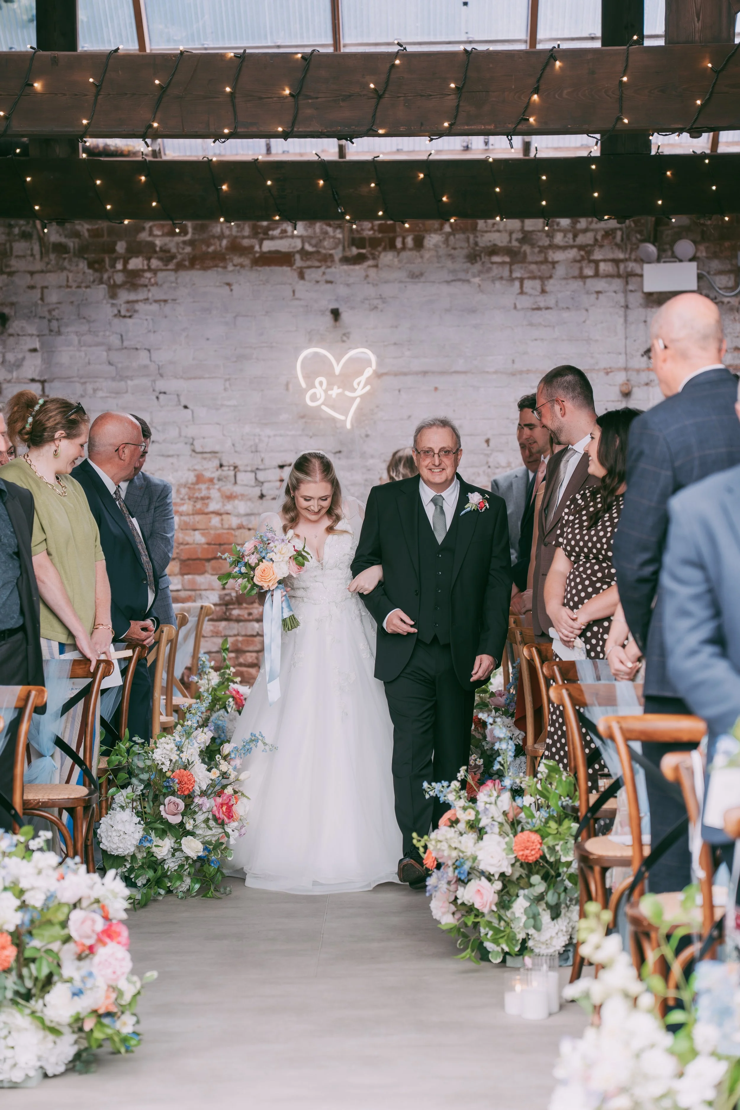 A bride and an older man, presumably her father, walking down the aisle at a wedding ceremony. The aisle is decorated with colorful flowers, and guests are standing on either side, smiling and watching. The background features a white painted brick w