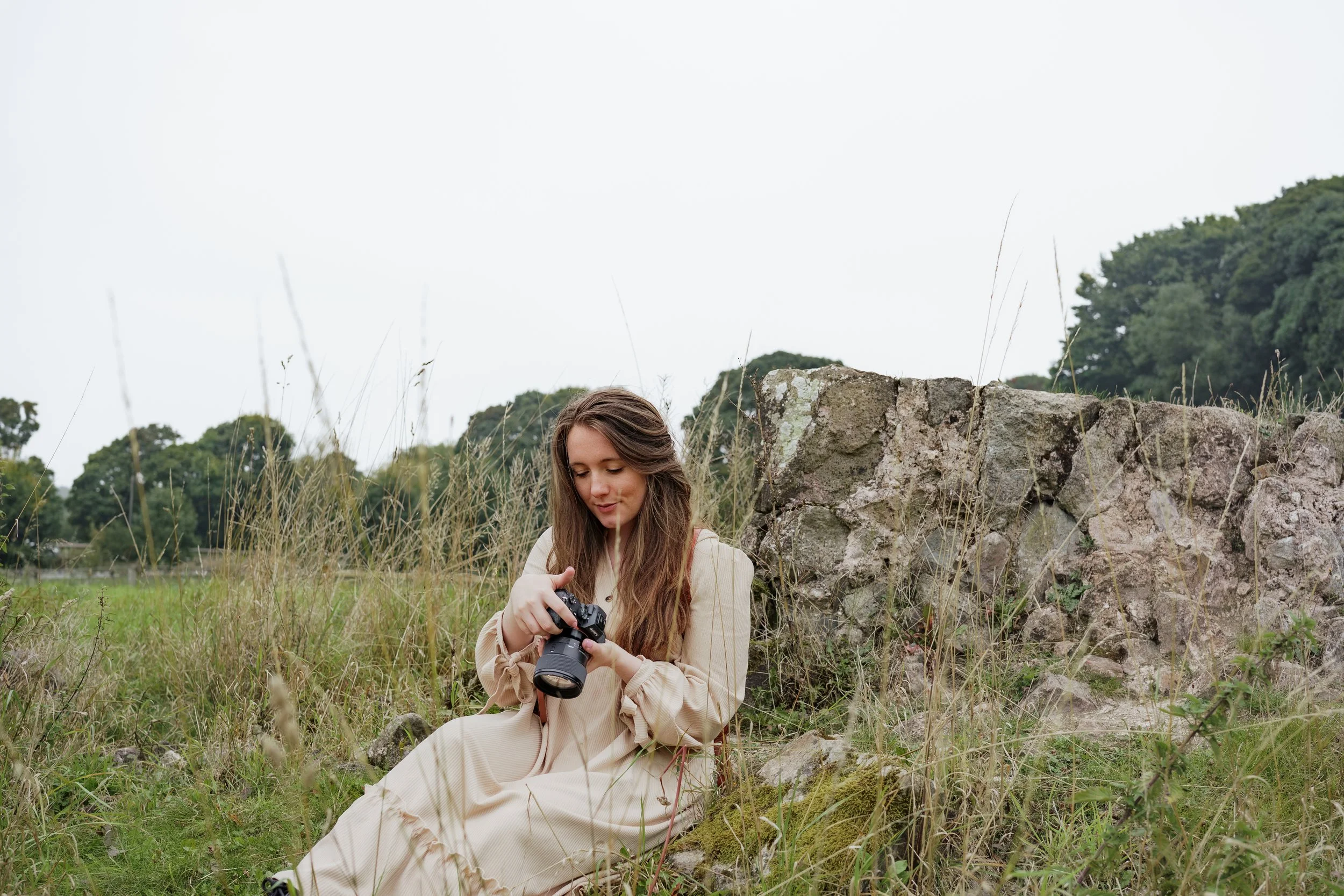 A young woman with long brown hair sitting outdoors near a stone wall, looking at her camera amid tall grass and trees in the background.