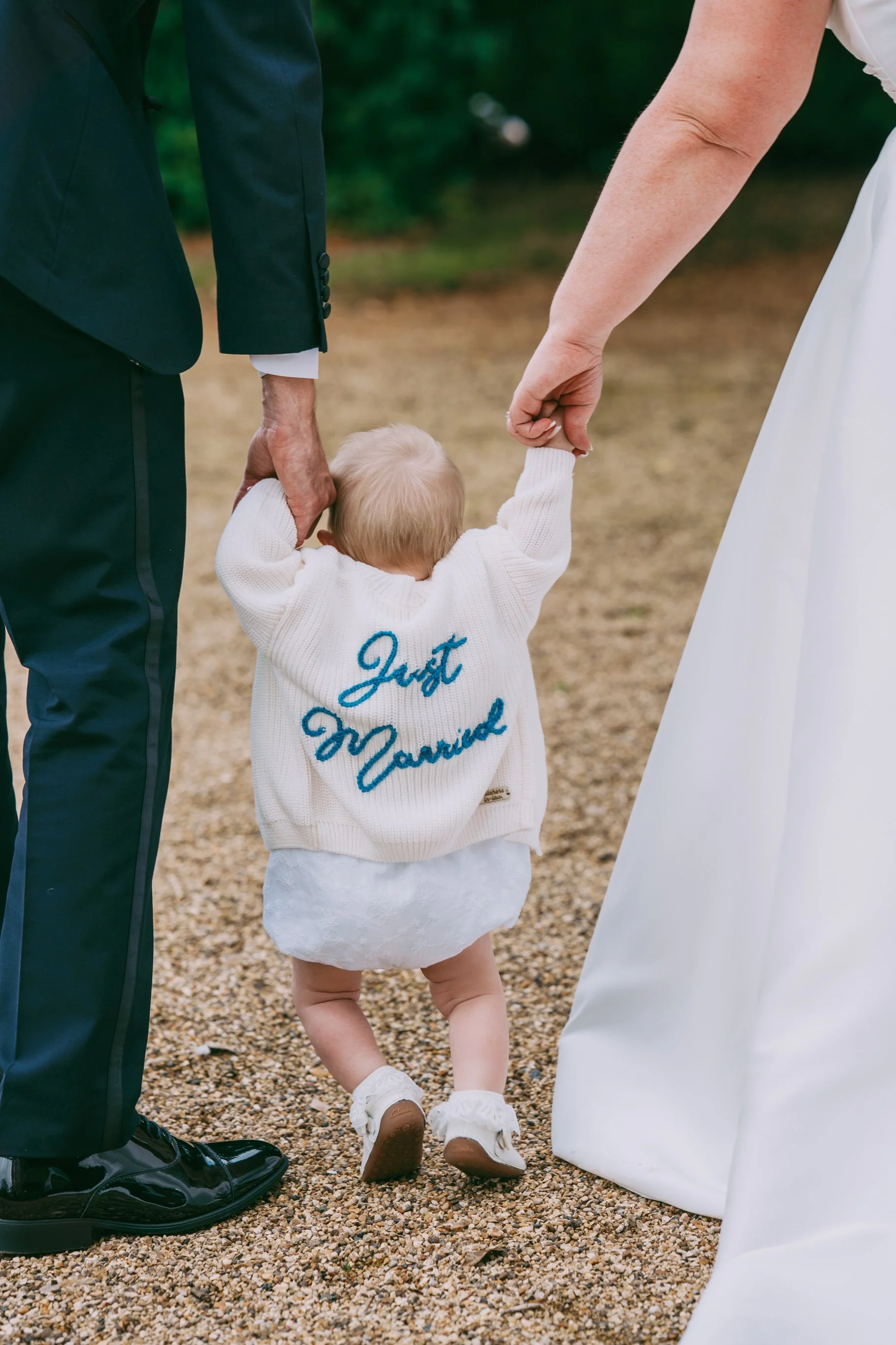 A toddler with blonde hair wearing a white sweater with blue embroidery holding hands with a man in a suit and a woman in a wedding dress during a wedding ceremony outdoors.