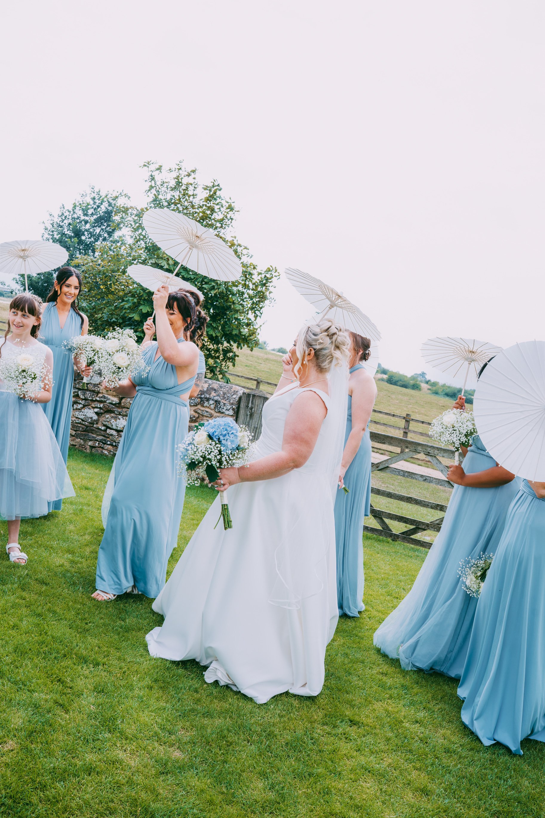 A group of women, some holding parasols and bouquets, outdoors on a grassy field during daytime, dressed in light blue and white dresses, possibly at a wedding or celebration.