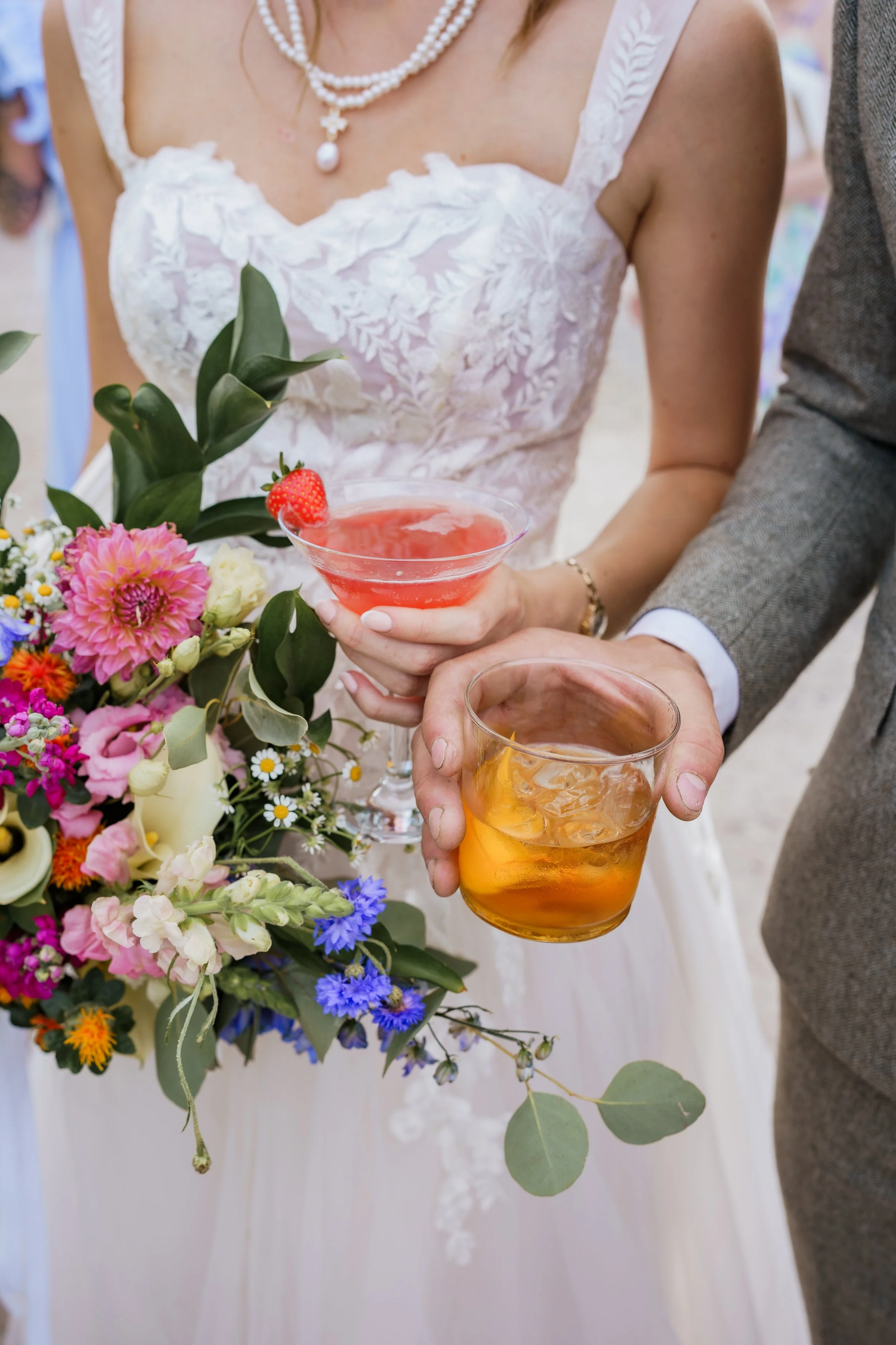 A bride holding a pink cocktail with a strawberry garnish, standing near a groom holding an amber-colored drink, with a bouquet of vibrant flowers and greenery nearby.