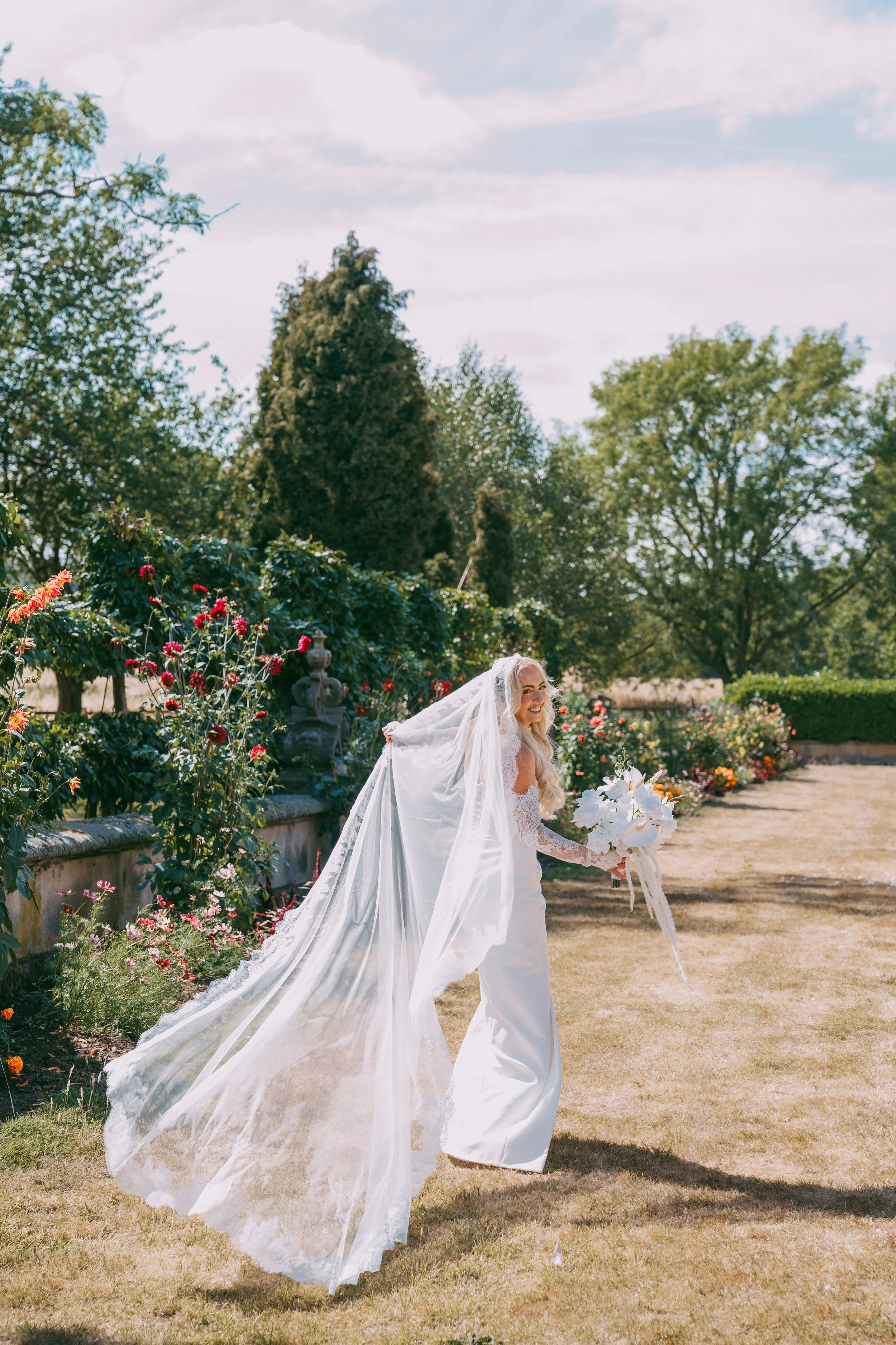 A bride standing outdoors in a garden with flowers, trees, and a partly cloudy sky, holding a bouquet of white flowers and wearing a white wedding gown with a long veil.