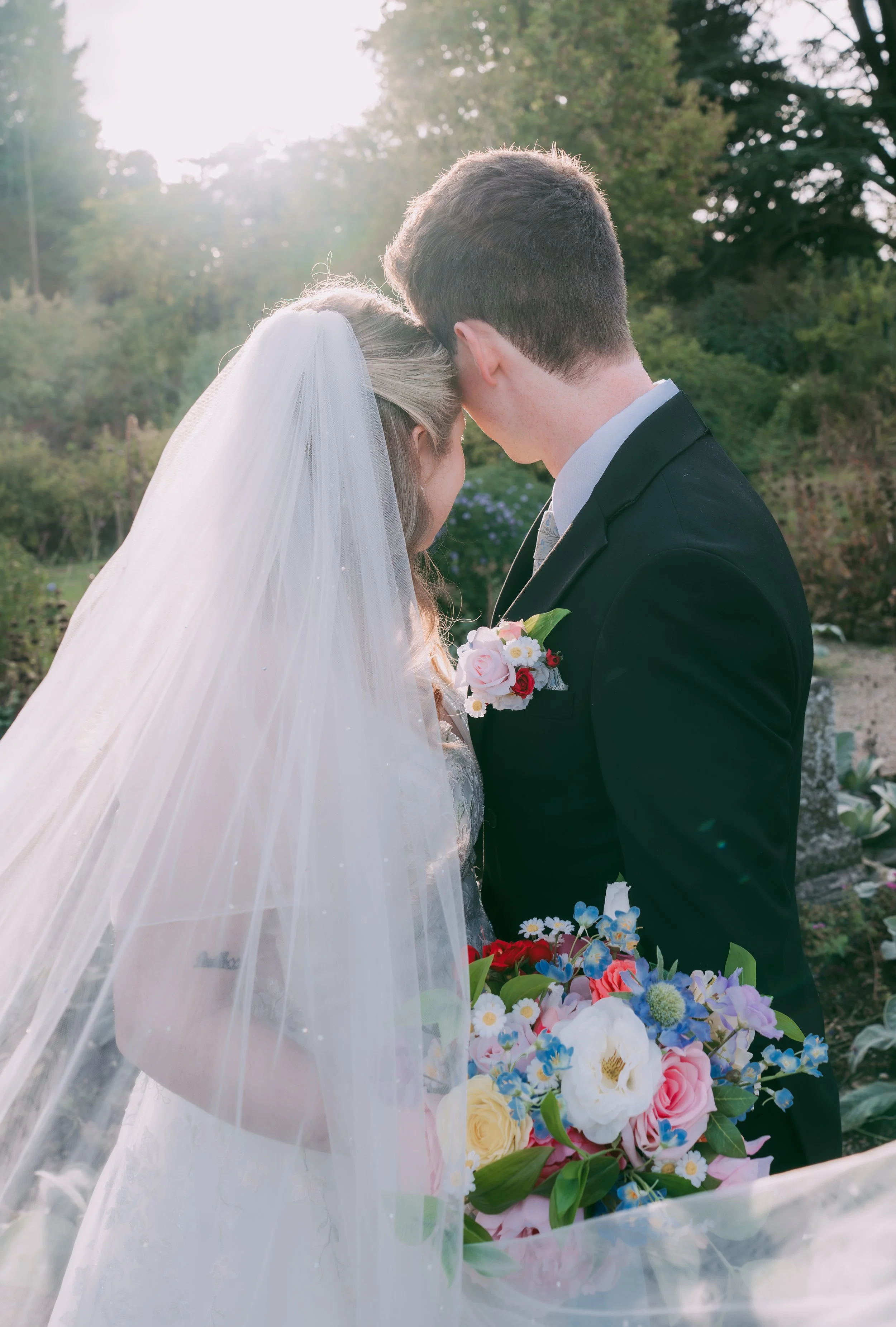 A bride and groom share a kiss outdoors during sunset, with the bride wearing a white wedding dress and veil, and the groom in a black suit, surrounded by greenery and holding a colorful flower bouquet.