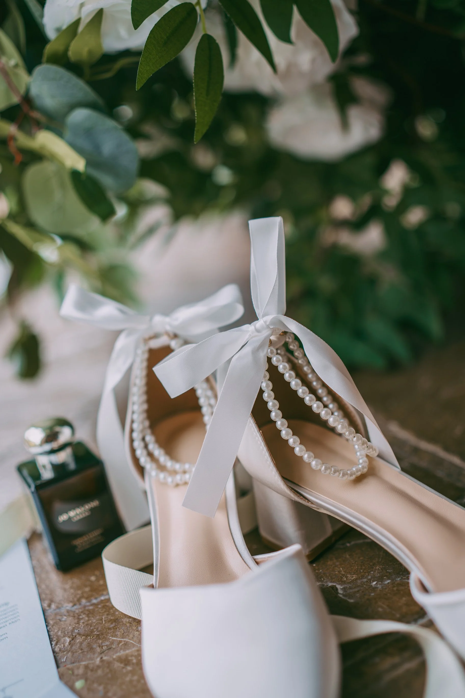 White satin high-heeled shoes with white ribbons, pearl necklaces, and perfume bottles on a wooden surface, with green foliage in the background.
