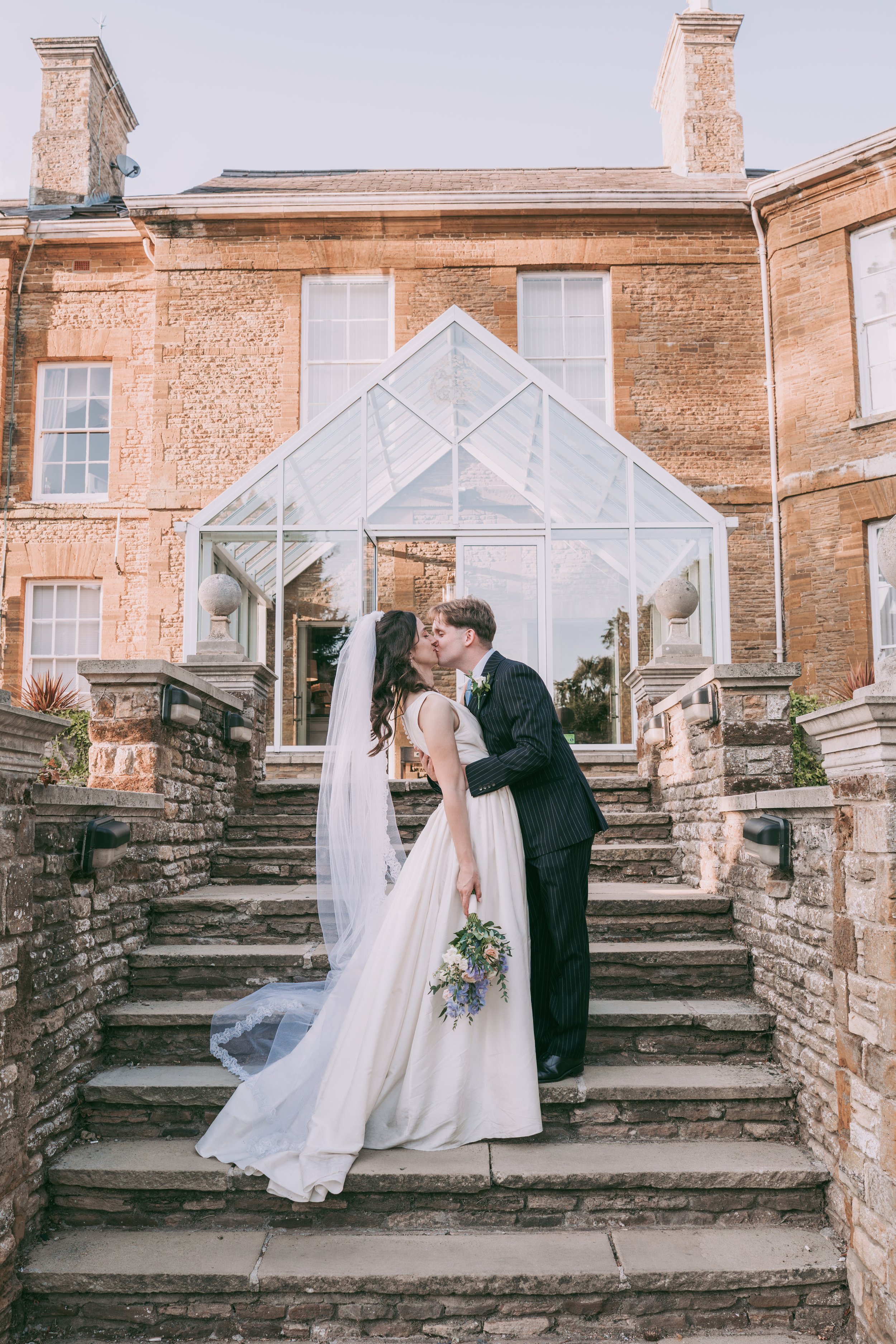 A bride and groom kissing on an outdoor stone staircase in front of a brick building with a glass conservatory.