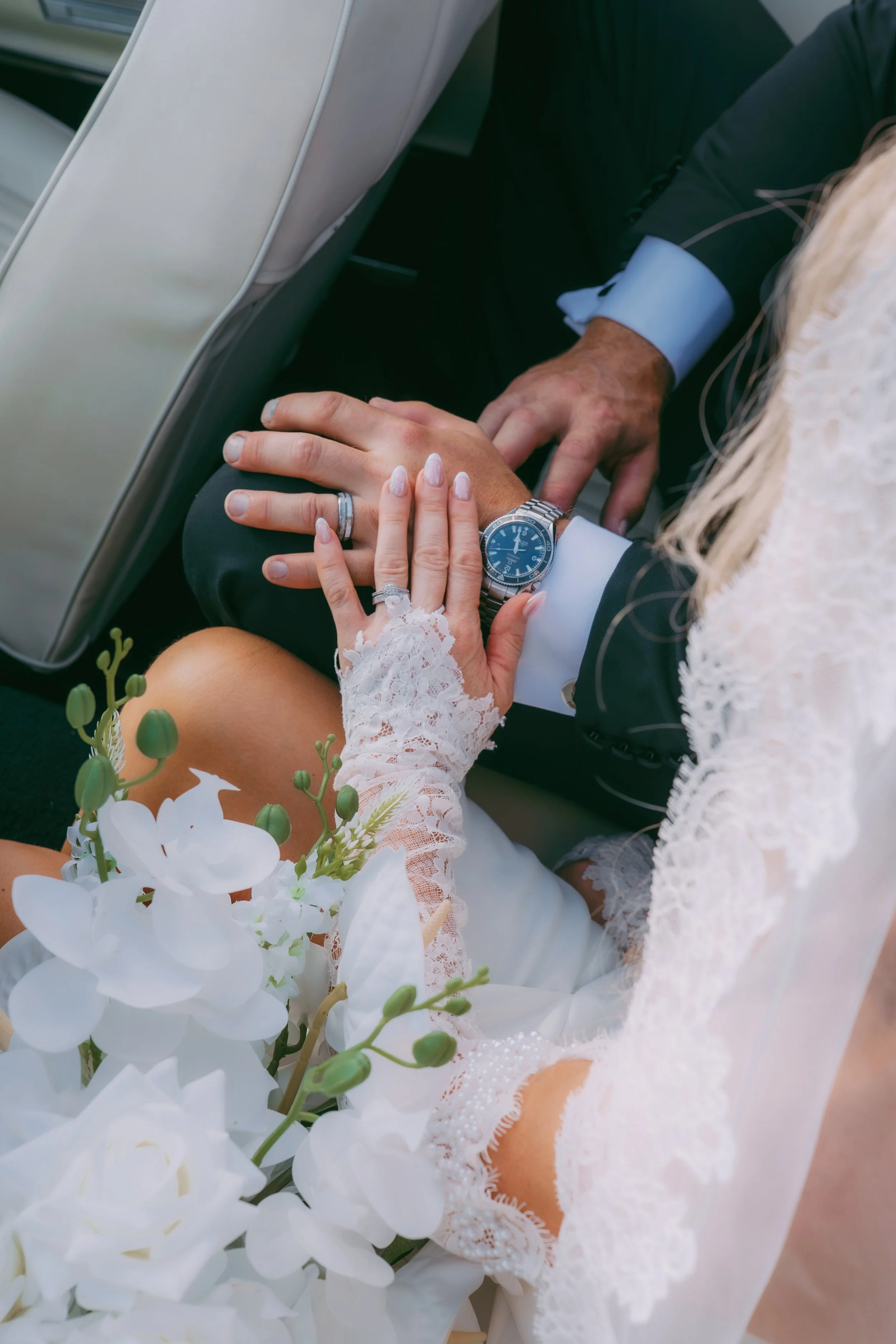 Close-up of a wedding couple's hands with rings, holding a bouquet of white orchids, with the bride wearing lace gloves and the groom wearing a watch, in a vehicle.