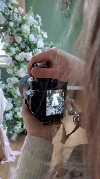 Person holding a camera taking a photo of a floral display with white, pink, and green flowers under a canopy