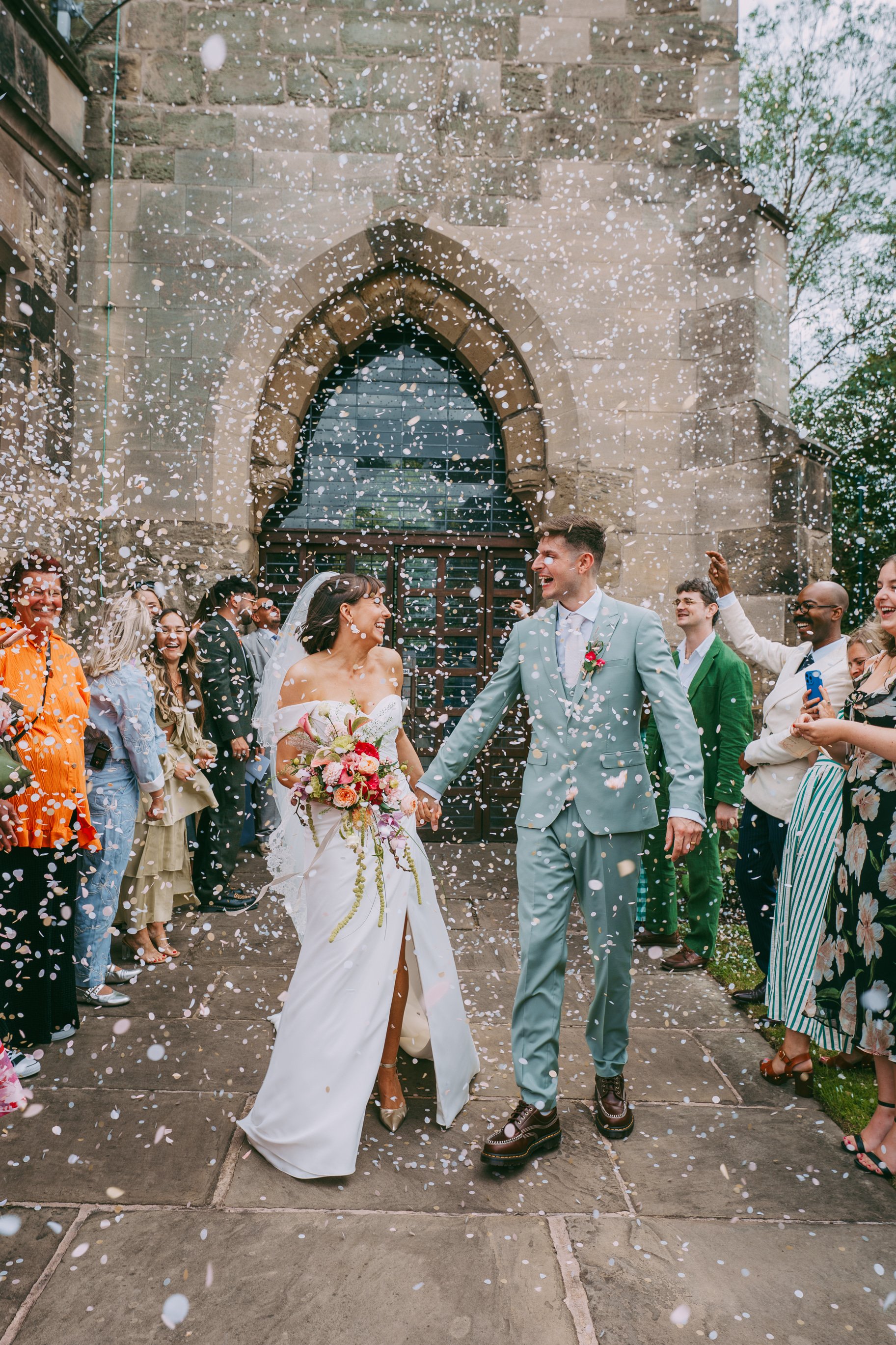 A newlywed couple holding hands, smiling, and surrounded by friends and family outside a stone church with confetti in the air, celebrating their wedding.