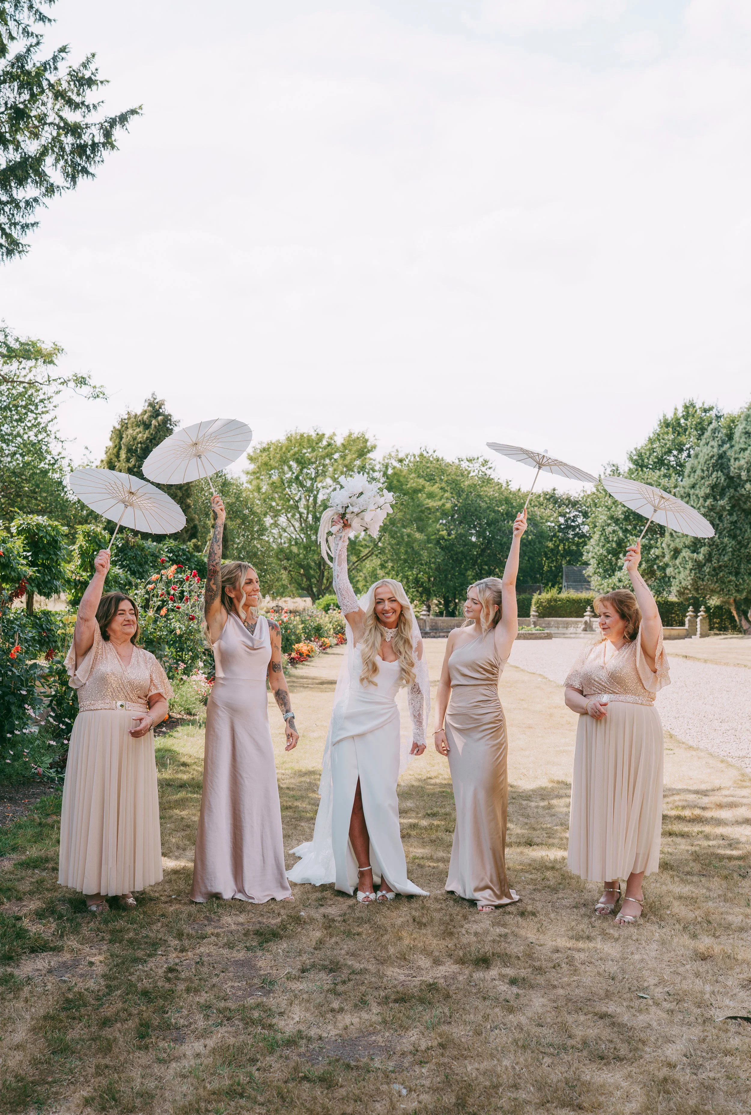 A group of women in elegant dresses under parasols outdoors, celebrating a wedding, with trees and a lawn in the background.