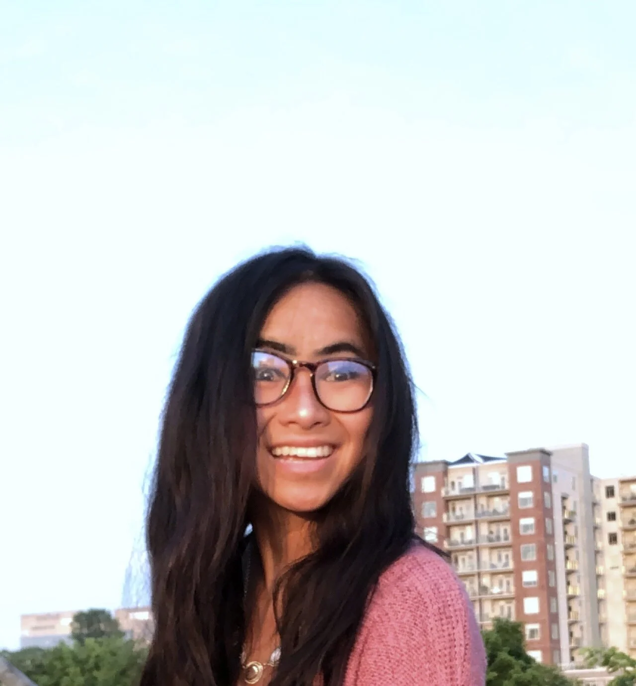 Smiling woman with long dark hair and glasses, outdoors in an urban area with buildings in the background during sunset.