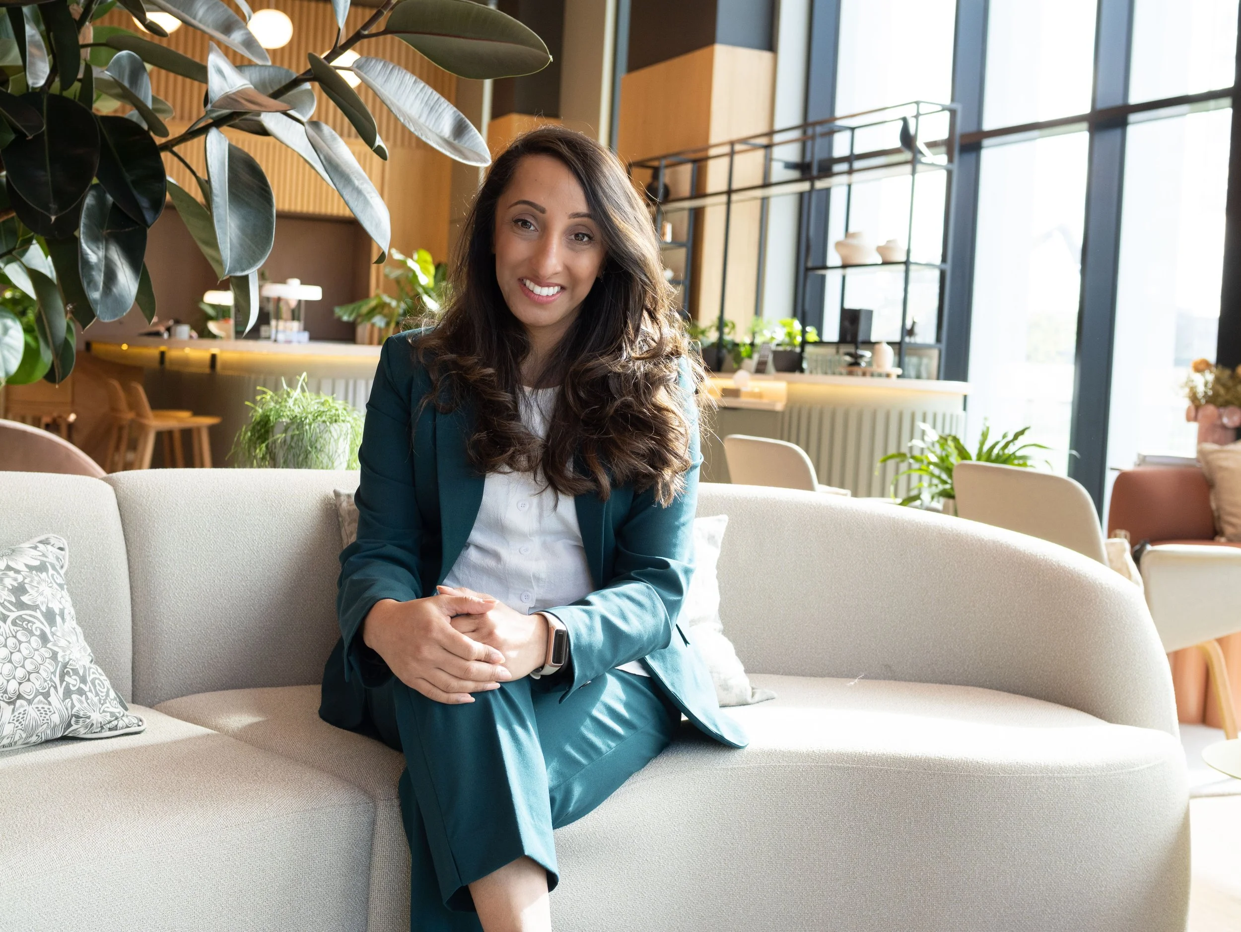 A woman with long curly brown hair, wearing a teal blazer and pants, sitting on a white sofa in a modern, well-lit indoor space with large windows, plants, and contemporary decor.