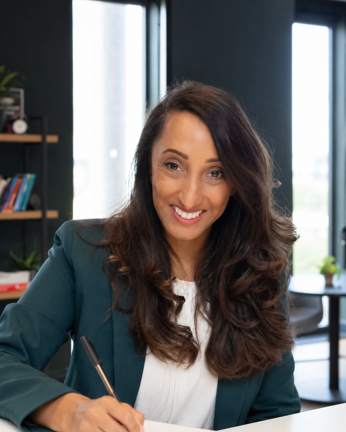A woman smiling and writing with a pen in a modern office with bookshelves and large windows in the background.