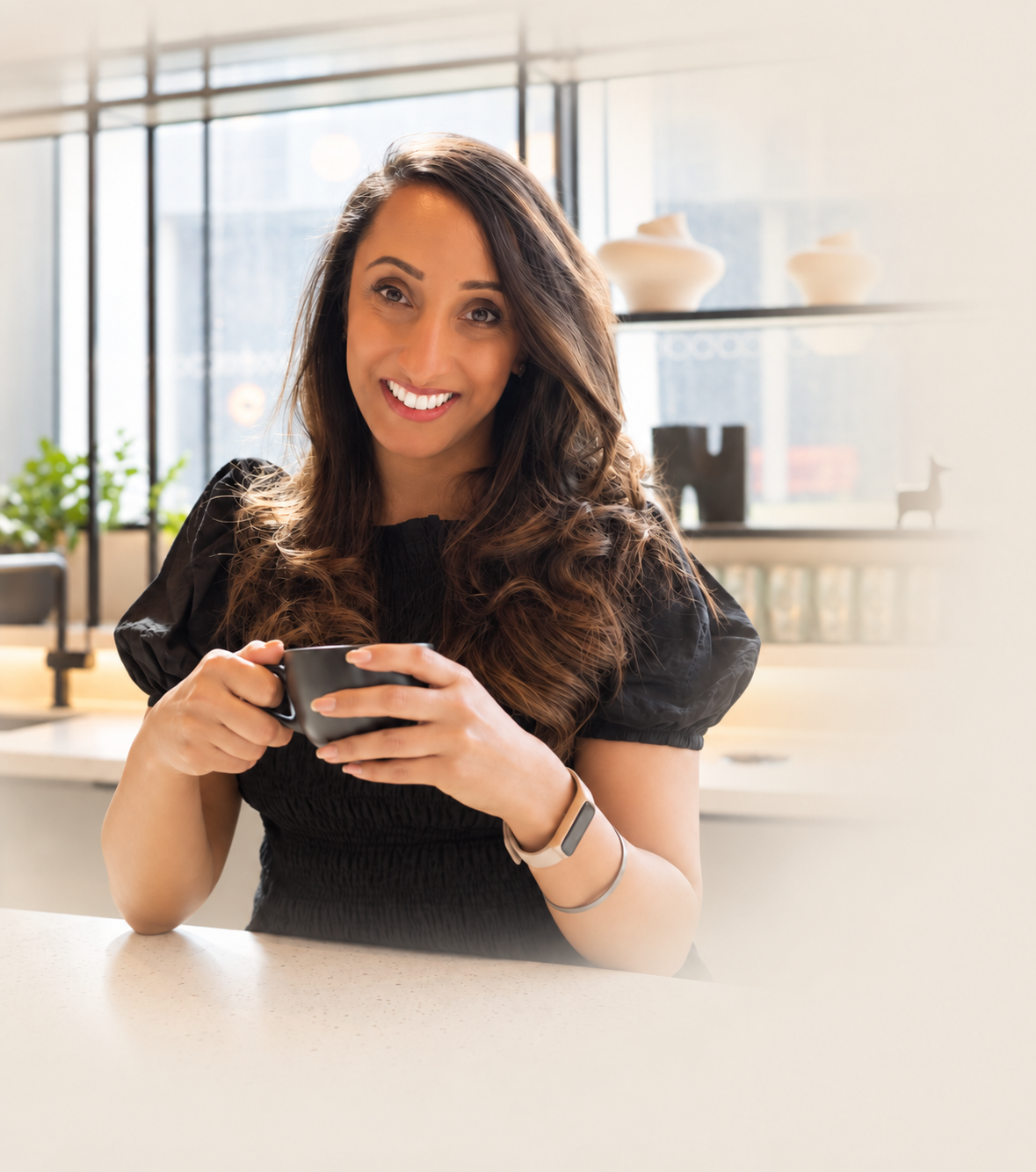 A woman with long brown hair smiling and holding a black mug in a bright modern kitchen.
