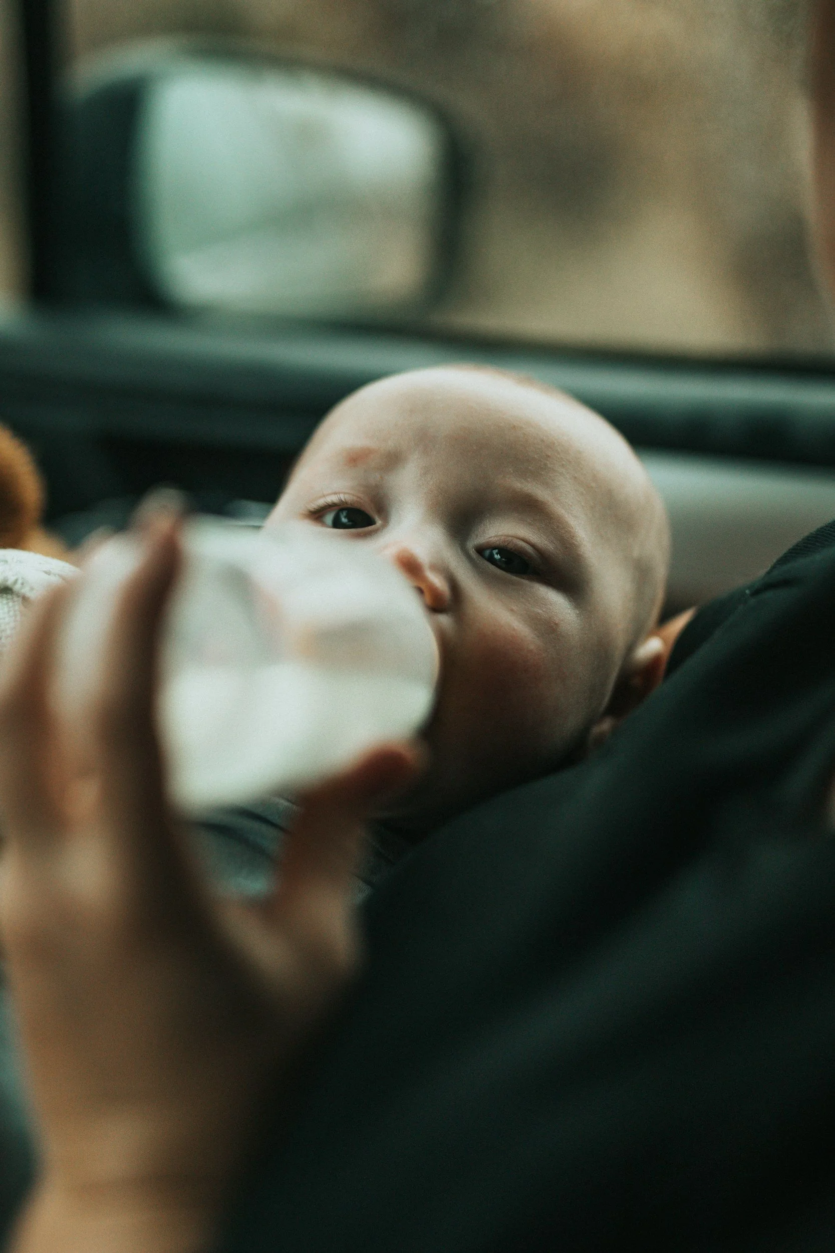 A baby with light skin and dark eyes looking up while drinking from a bottle, held by someone outside the frame.