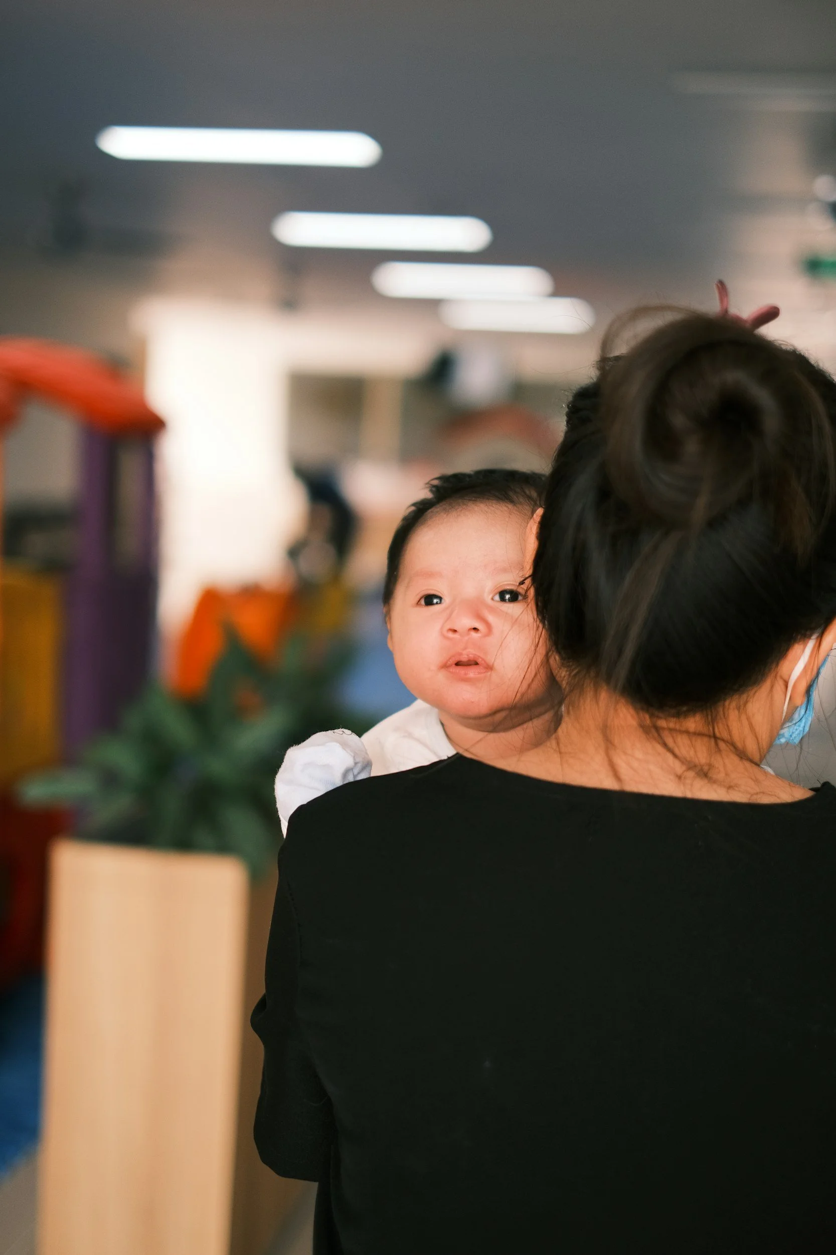 A woman with dark hair tied up, wearing a face mask, holding a small baby with big eyes, indoors with blurry ceiling lights in the background.