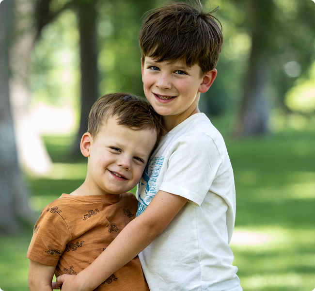 Two young boys hugging outdoors in a park with green grass and trees in the background, smiling at the camera.