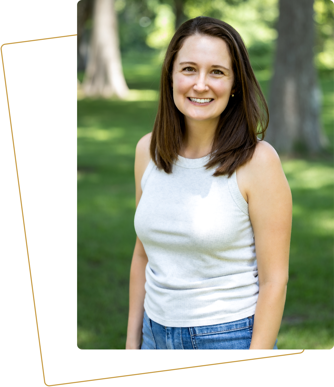 Smiling woman standing outdoors with trees and green grass in the background.
