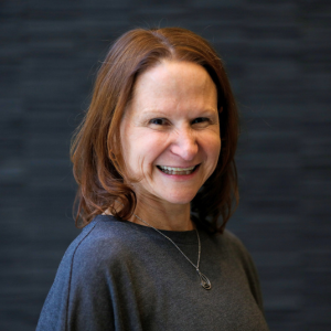 A woman with shoulder-length reddish-brown hair smiling, wearing a dark gray top and a necklace, standing against a dark background.