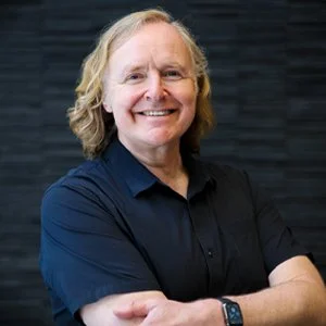 Smiling woman with shoulder-length blonde hair wearing a navy blue shirt, arms crossed, standing against a dark background.