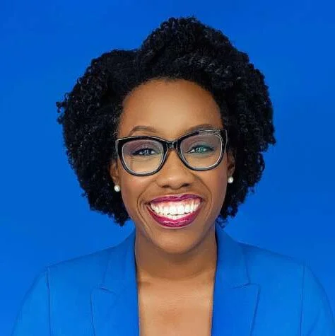 Close-up of a smiling African American woman with curly black hair, glasses, and pearl earrings, wearing a blue blazer against a blue background.