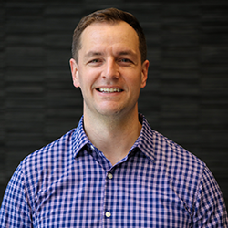 Headshot of a smiling man wearing a blue checkered shirt against a dark textured background.