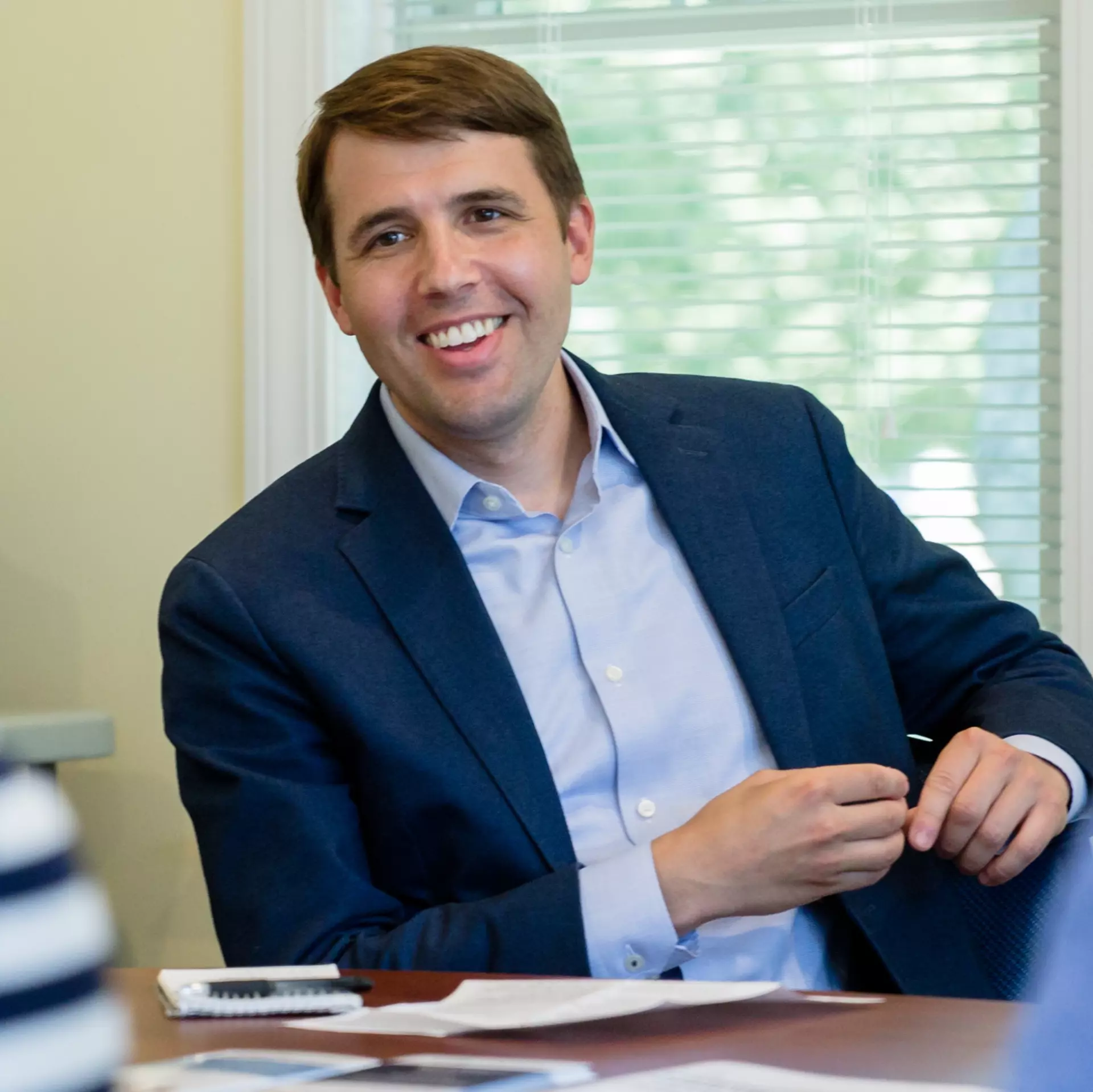 Smiling man in a business suit sitting at a desk with papers, a calculator, and a pen, in a room with window blinds.