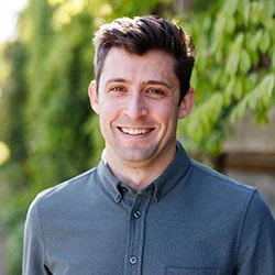 A young man with dark hair and a friendly smile, wearing a button-up shirt, outdoors with green trees in the background.